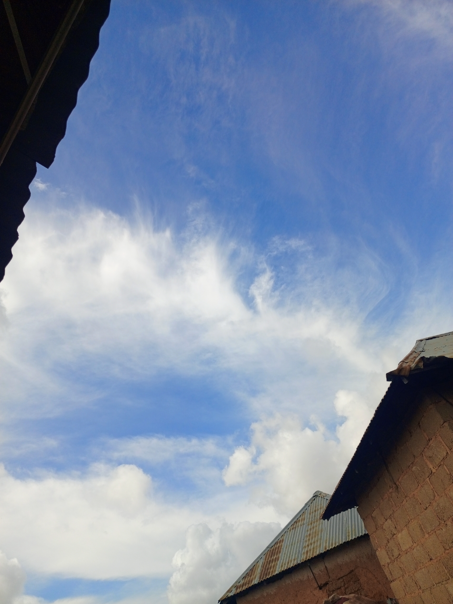 This is a low-angle, full-shot photograph of a bright blue sky filled with wispy white clouds. In the foreground, the roofs of two buildings are visible. The building on the left is mostly obscured by a dark, textured overhang, possibly part of a corrugated metal roof. The building on the right is constructed from reddish-brown mud bricks, and its corrugated metal roof is visible at an angle. The scene appears to be outdoors in a residential area, potentially in Jos, Nigeria, based on the architectural style and materials used. There are no people or animals visible in the image, and no discernible actions or interactions are taking place. The environment is characterized by clear weather conditions with scattered clouds.