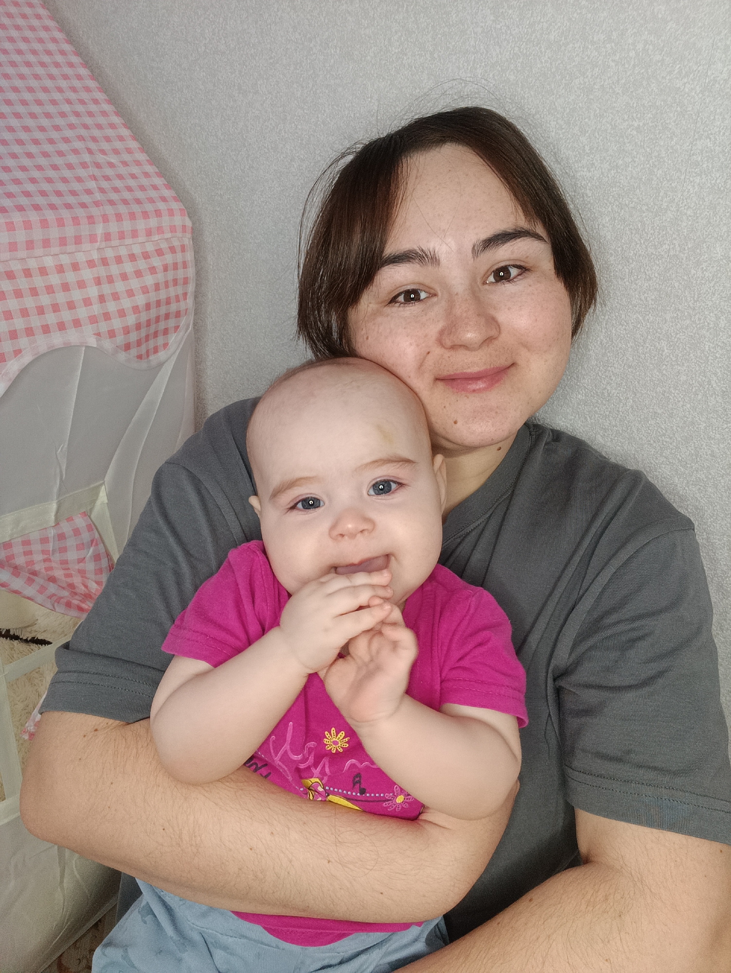 A woman holds a baby in her arms. The woman has short brown hair, freckles, and is wearing a gray shirt. The baby is wearing a pink shirt and blue pants, with a small bruise on her forehead. The baby has blue eyes and is holding her hands to her mouth. They are indoors, in front of a light gray wall and next to a white crib with a pink and white checkered pattern. The location in the city of Noviy, Russia, cannot be determined from the image.