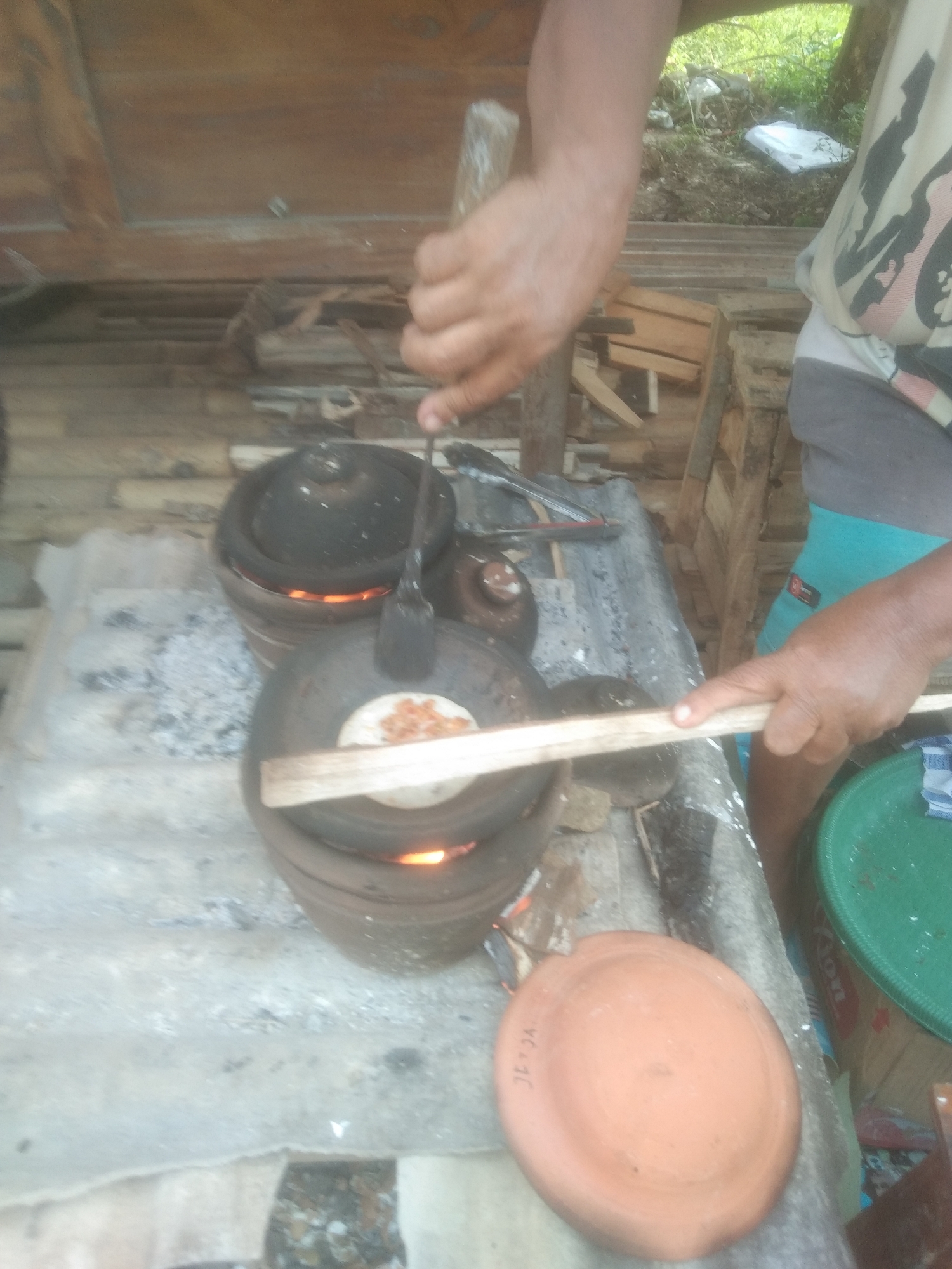 A person is cooking outdoors, likely on a makeshift stove, as indicated by the presence of ash and flames visible beneath two clay pots. The person is wearing a t-shirt with a camouflage pattern and teal shorts. They are holding a wooden stick, possibly to stoke the fire or manipulate the cooking food. One of the clay pots has a flat lid with a piece of flatbread and some toppings cooking on it. A wooden plank is placed across the pot, suggesting it is being used as a makeshift tool. In the foreground, there is a terracotta-colored plate with some markings. The background shows a rustic wooden structure and some greenery, suggesting a rural or semi-rural setting. The image's quality is grainy, making precise details difficult to discern. This scene is characteristic of informal food preparation often found in local markets or homes in areas like Subang, Indonesia, where traditional cooking methods are still prevalent.
