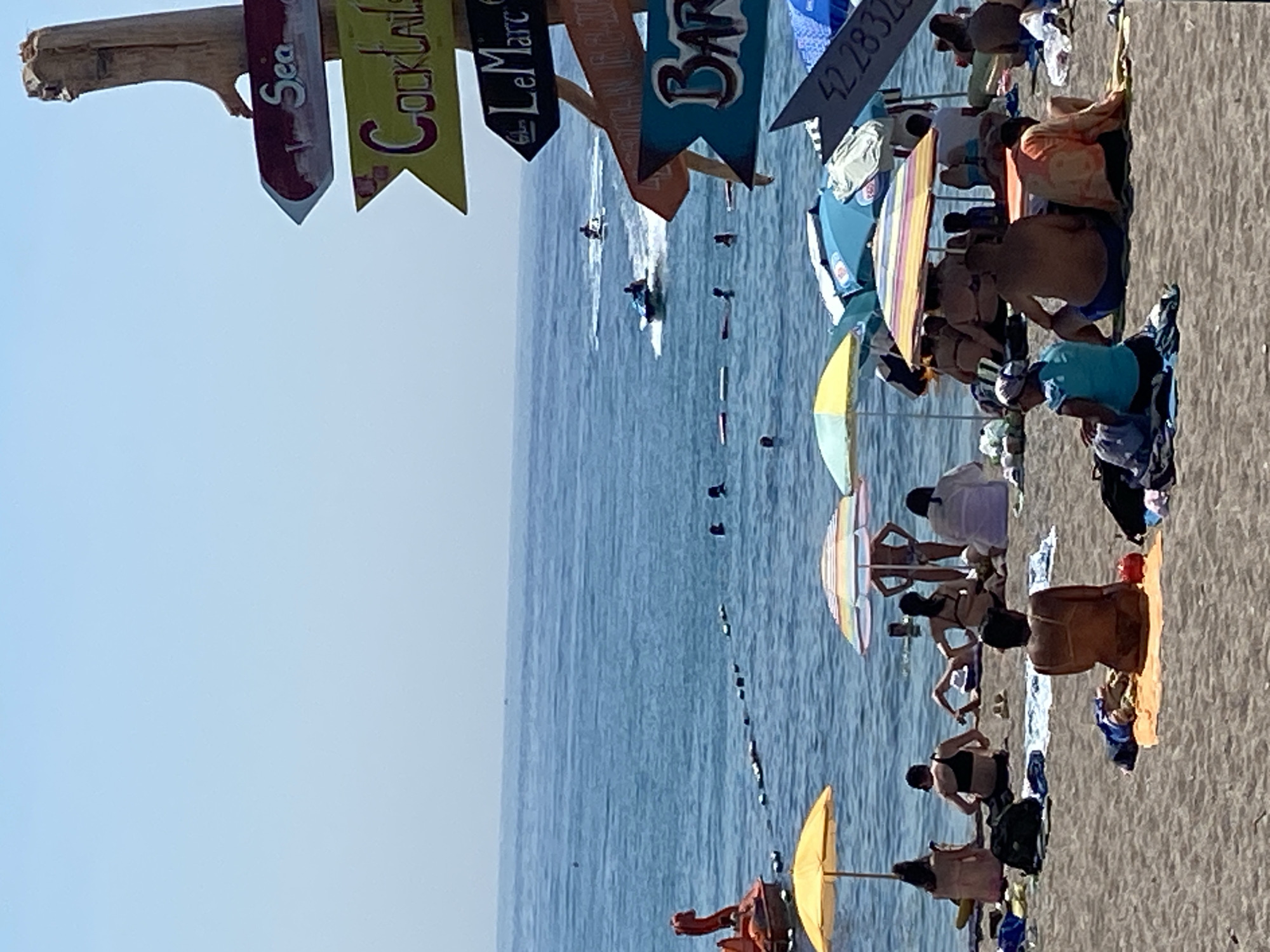 This is an eye-level, medium shot of a crowded beach in Prijevor, Montenegro on a clear, sunny day. The foreground is dominated by a sandy beach populated by numerous beachgoers, many of whom are seated or reclining on towels and under colorful umbrellas. In the mid-ground, the calm, blue sea stretches to the horizon, dotted with a few distant jet skiers and swimmers. A red swan-shaped watercraft is visible on the left side of the frame. On the right side of the image, a wooden post with several directional signs points towards "Sea", "Cocktails", "Le Marc", and "Bar". One sign also displays a numerical sequence, "42 2832". The overall atmosphere is one of leisure and recreation, typical of a summer beach scene.