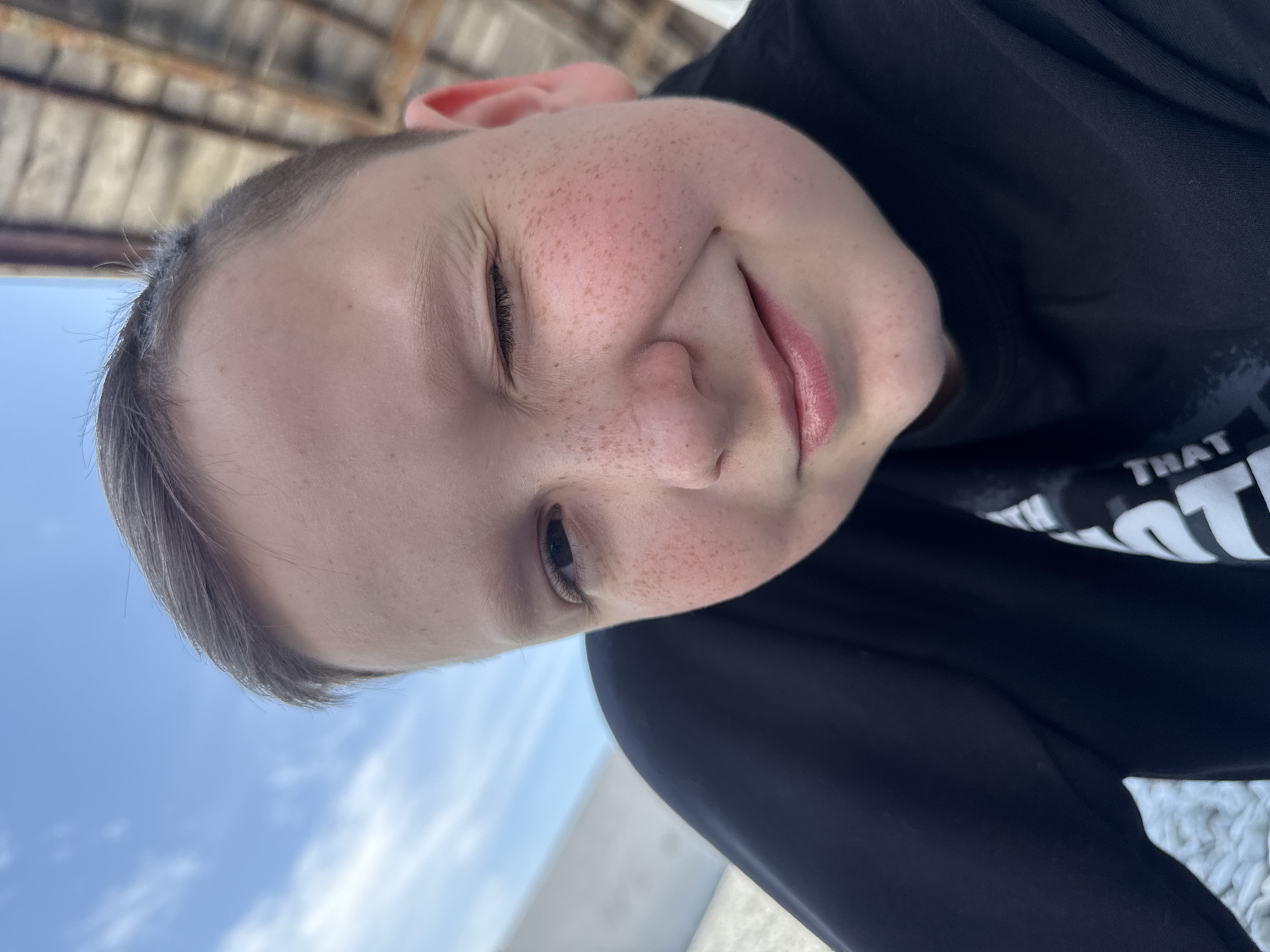 A portrait of a fair-skinned child with freckles, likely a boy, is captured from a low angle, with the camera tilted sideways. The child has short, light brown hair. The background includes a bright blue sky with soft, white clouds and a blurry wooden structure. The child is wearing a black t-shirt. The location is unknown.