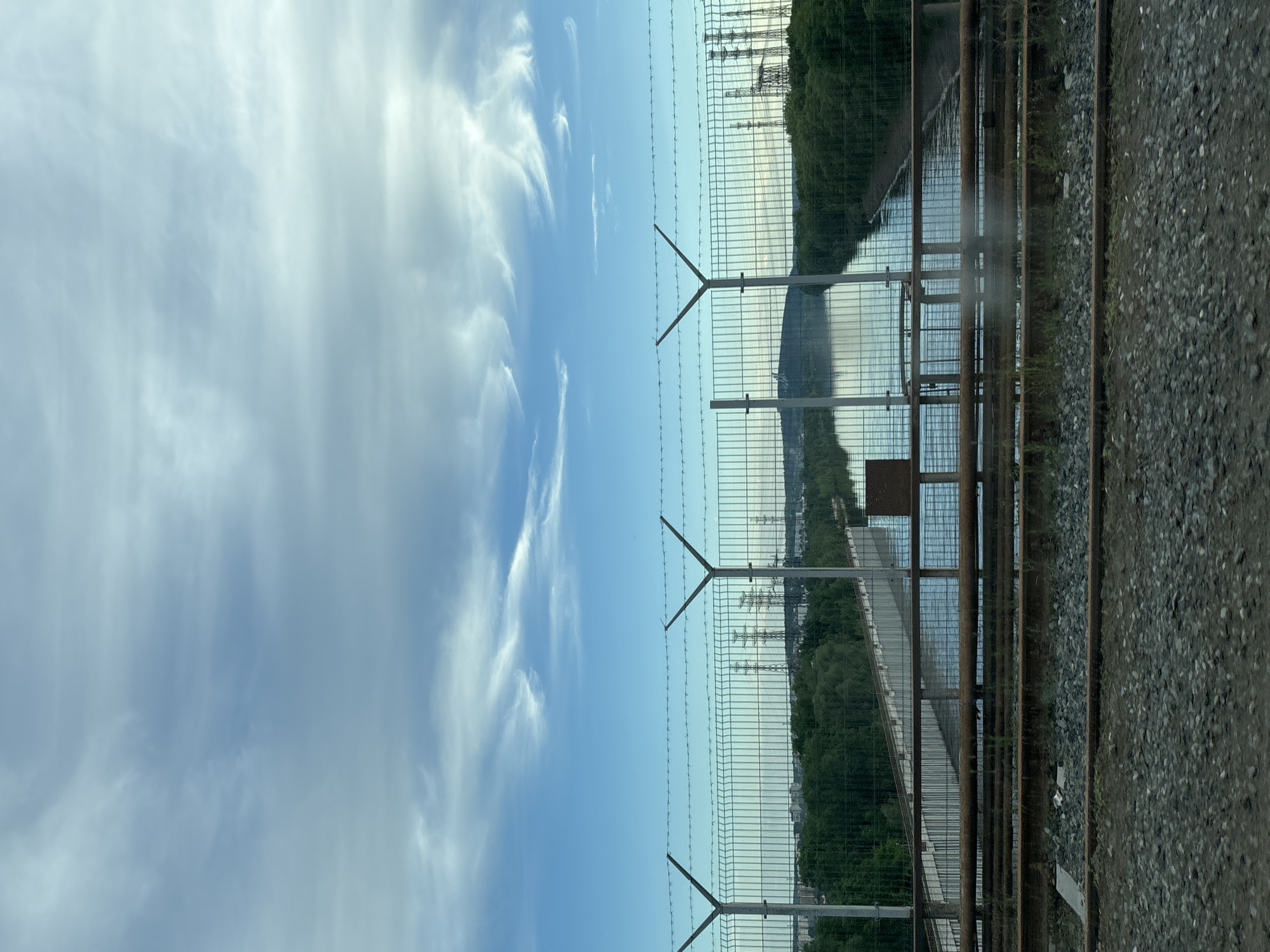 The image shows a view through a wire fence with barbed wire at the top. Beyond the fence is a body of water, possibly a river or reservoir. Trees line the banks of the water. Power lines are visible in the distance. To the right of the fence is a gravel area, likely a railway or industrial zone. The sky is blue with some clouds. Based on the clues, this image is possibly located in Chaikovsky, Russia.
