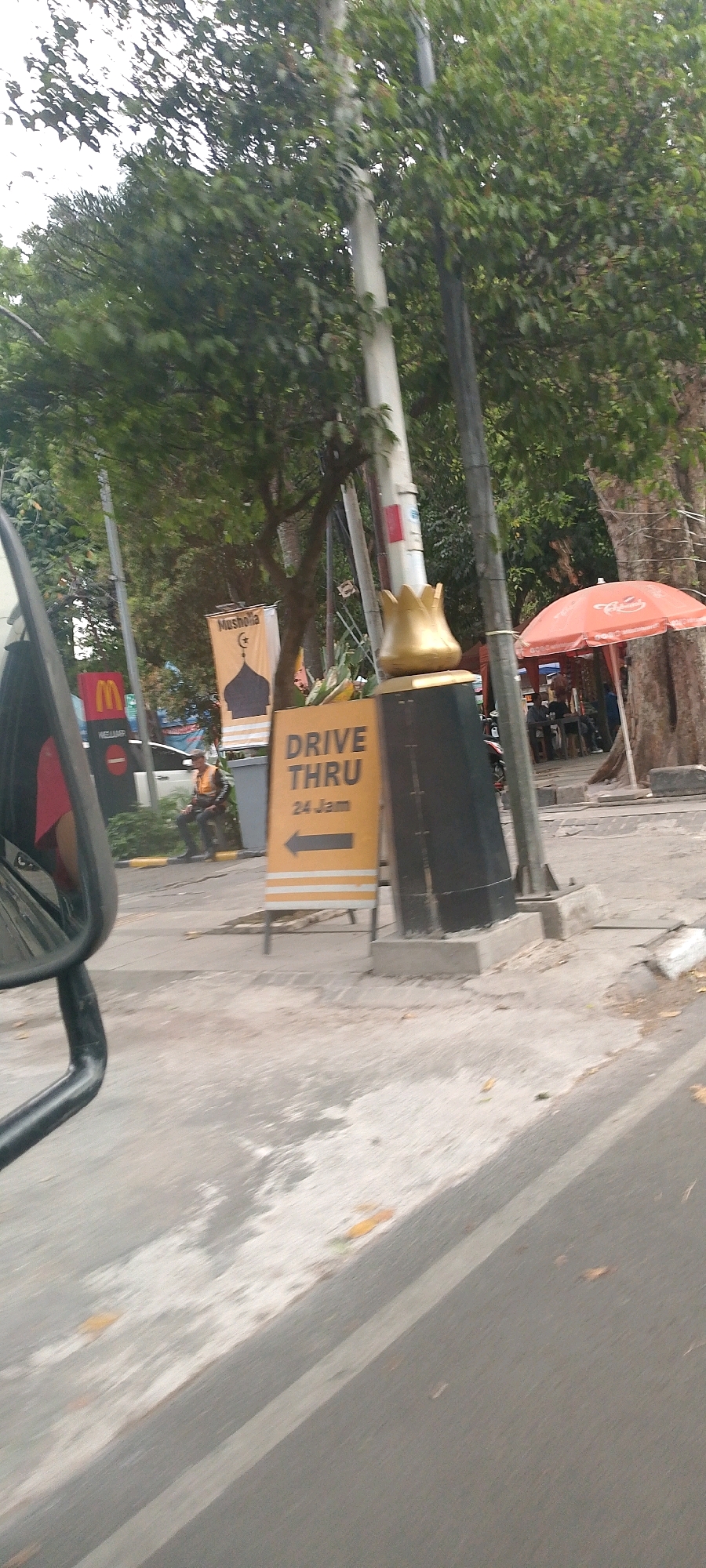 The image captures a street scene in Bandung City, Indonesia, likely in front of a McDonald's establishment. In the foreground, a side mirror of a vehicle is visible on the left, suggesting the photo is taken from a moving car. A yellow sign with black text that reads "DRIVE THRU 24 Jam" (24 Hours) is prominently displayed on a black pedestal. To the left of this sign, a banner featuring a mosque silhouette and Islamic script is hanging, accompanied by the word "Musholla". A red umbrella, possibly part of a food stall or outdoor seating area, is visible in the background on the right. A person in a high-visibility vest is seated on a low wall in the middle ground, near a white vehicle. Trees and foliage line the background, creating a somewhat shaded environment. The overall scene depicts a typical urban commercial area with elements of both fast food and religious services.
