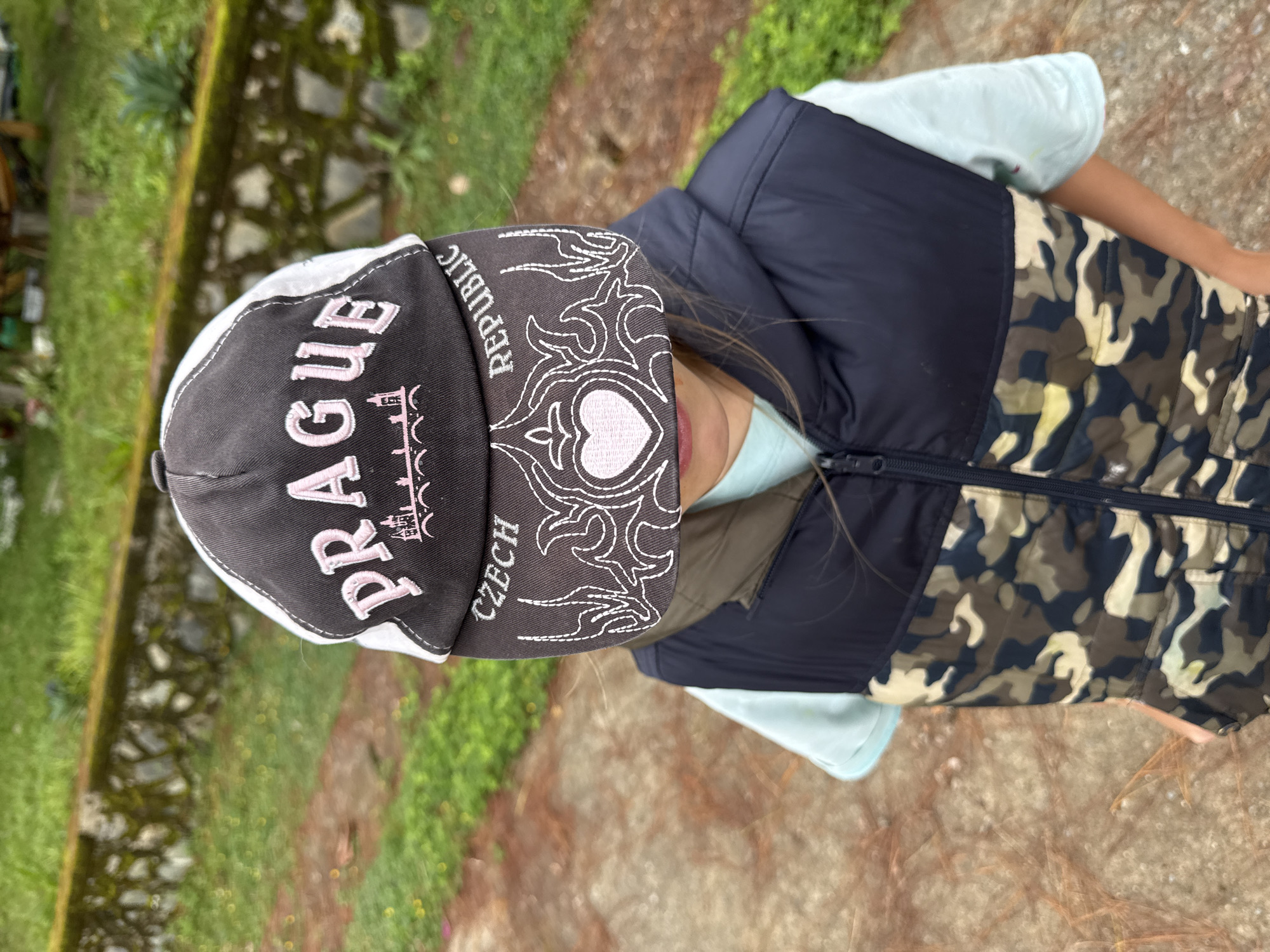 A young person wearing a baseball cap and a vest stands outside on a path. The cap is black and white with "PRAGUE CZECH REPUBLIC" written on it in pink and white. The vest is camouflage with a black top. The path is made of concrete and has a dirt and leaf-covered area next to it. Behind the path is a stone wall and lush green grass. The image was taken in the city of Đạ Cháy, Vietnam.