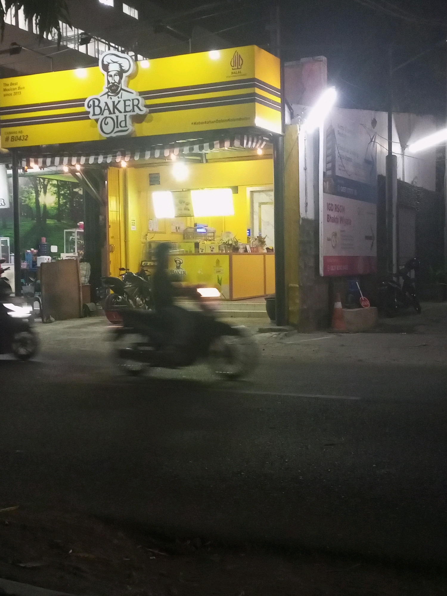 The image shows a street scene at night in Kediri City, Indonesia. A yellow storefront with a black awning and string lights displays a sign that reads "BAKER OLD" with a stylized chef logo. The text on the sign also indicates "The Best Mexican Bun since 2015" and "#B0432". To the right of the storefront, a sign for "IGD RSGM Bhakti Wiyata" is visible. The street in front of the establishment is dark and appears wet, with a blurred image of a motorcycle rider in motion. Other motorcycles are parked near the storefront. The overall atmosphere is of a commercial area at night.