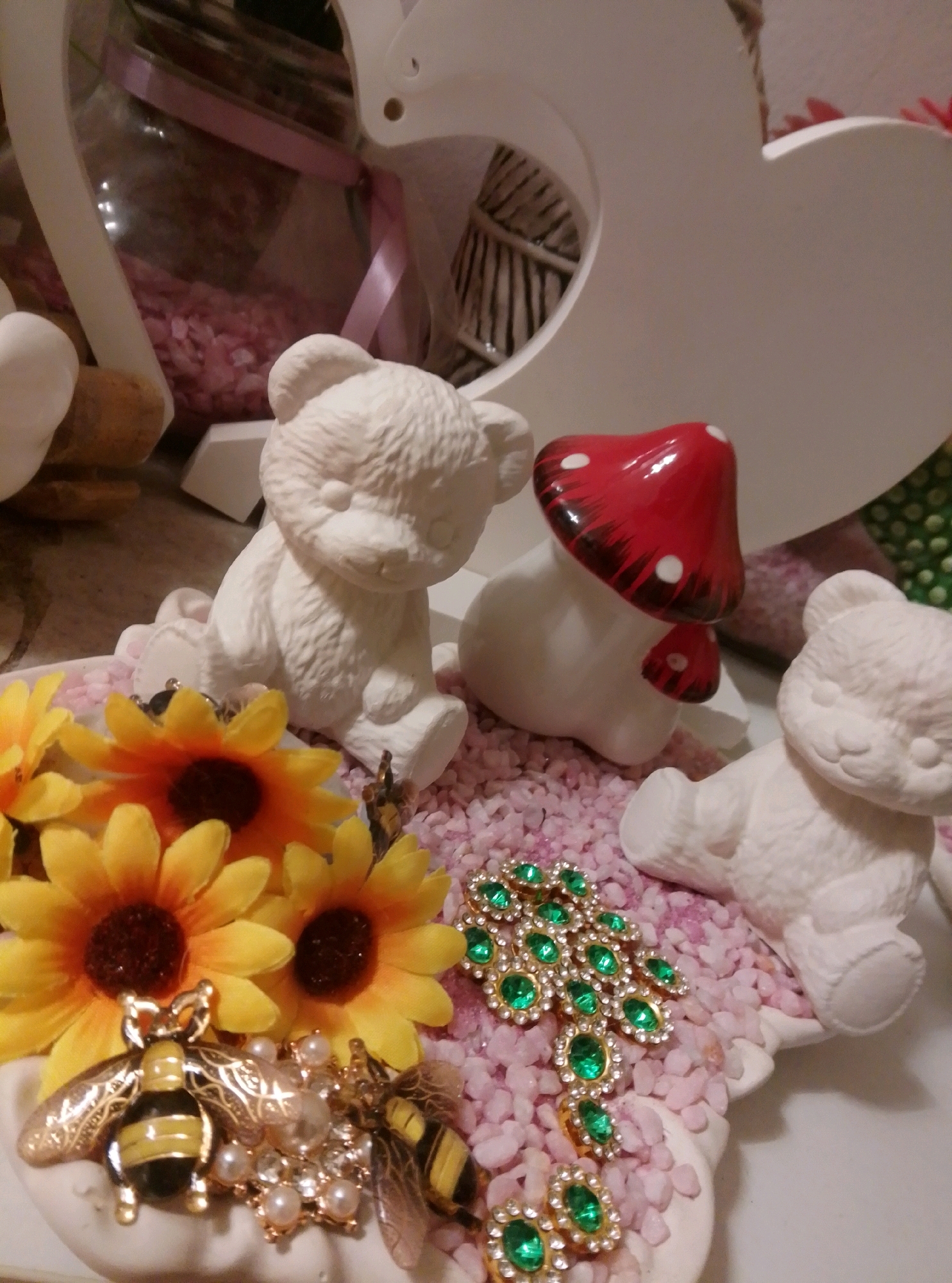 This is a close-up, eye-level, medium shot of a collection of decorative items arranged on a pink, pebbled surface. The scene is brightly lit, with a shallow depth of field. In the foreground, two gold and black bumblebee figurines are positioned next to a cluster of faux sunflowers. A gold brooch adorned with numerous green gemstones and clear beads is also visible. Behind these, two unpainted, white ceramic teddy bear figurines are seated, one on the left facing forward and another on the right facing slightly to the left. Between the teddy bears, a white ceramic mushroom figurine with a red cap decorated with white spots is placed. A large, white, heart-shaped cutout forms the background. To the far left, part of a glass jar containing pink pebbles and a pink ribbon is visible. The overall aesthetic is whimsical and decorative. There is no indication of the city of Wedel, Germany, in the image.