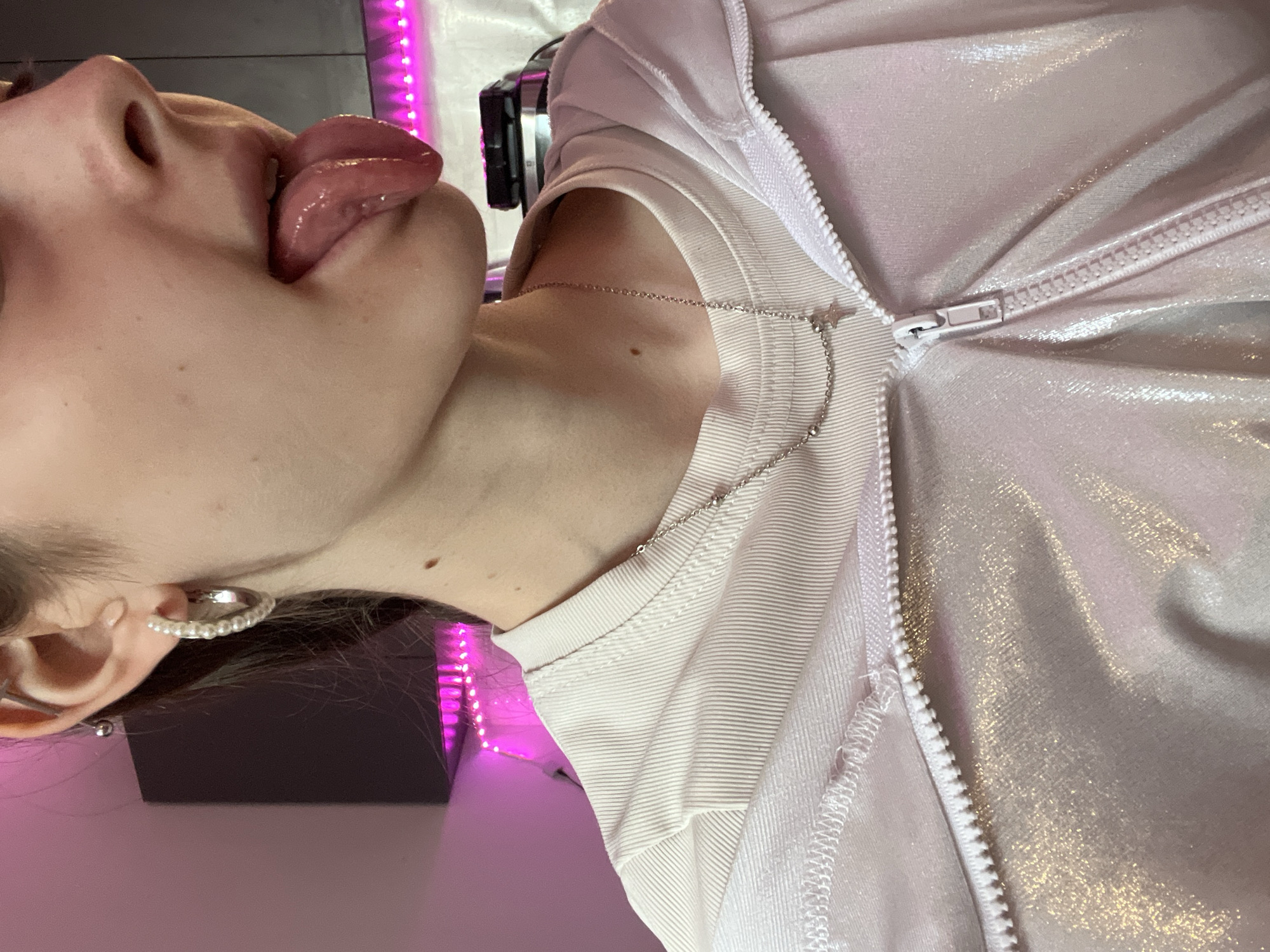 A close-up, sideways, studio shot shows a person with pale skin and light brown hair sticking their tongue out. The person is wearing a white t-shirt under a light pink, iridescent zip-up jacket with a white zipper. A silver chain necklace hangs around the neck. The person is wearing a silver hoop earring with small white beads. The background is a gray wall and a purple LED light strip.