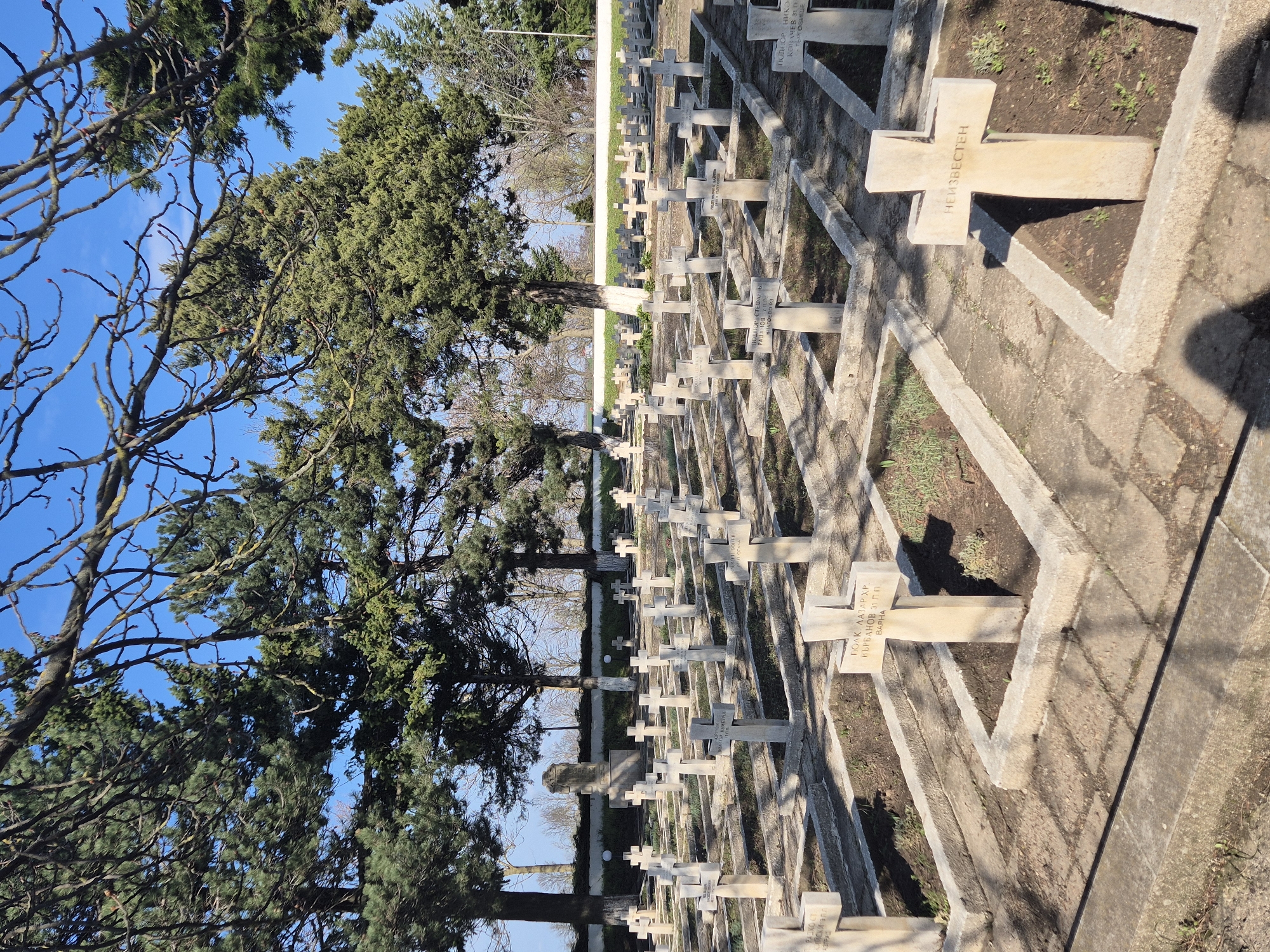The image shows a cemetery with rows of stone crosses under a blue sky and trees in Shumen, Bulgaria. The crosses bear inscriptions in Cyrillic script, including the names of the deceased. The setting suggests a location of remembrance and respect for the deceased. The crosses are set in concrete enclosures with patches of dirt and greenery. The overall impression is one of order and reverence.