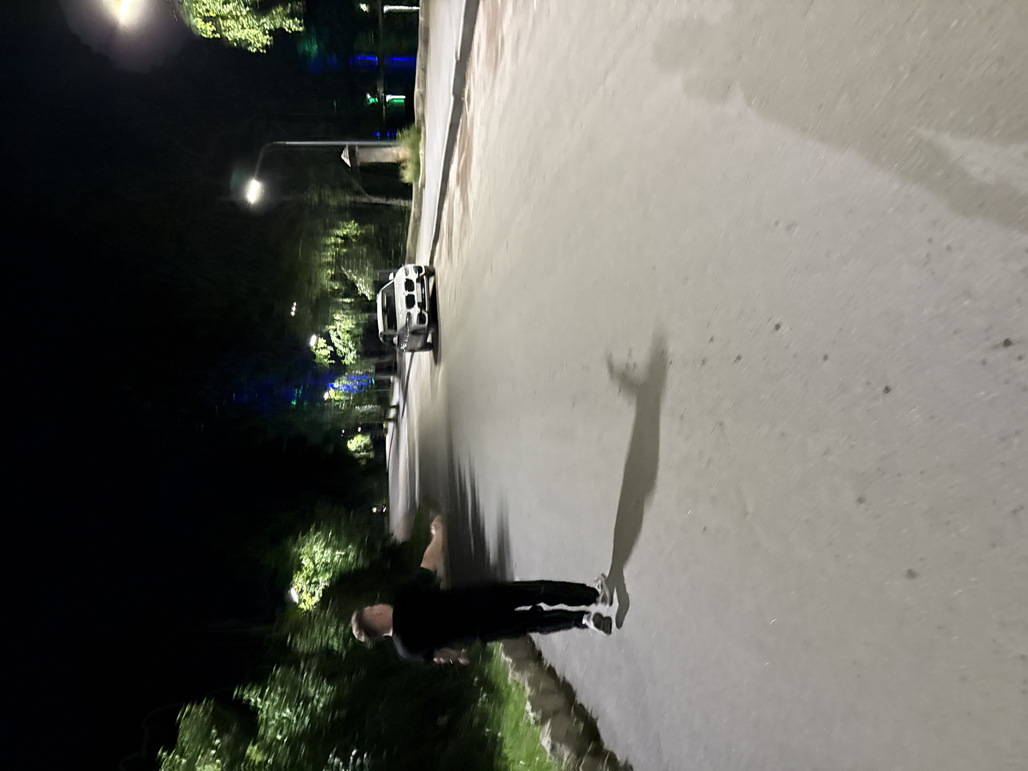 A man in a black t-shirt and pants is walking away from the camera on a paved road at night. He is in motion, with his arms slightly extended. To his right, further down the road, a white SUV is parked. The scene is lit by streetlights, casting long shadows. Trees and foliage are visible on the left side of the road, and a body of water with lights can be seen in the background on the right. The environment suggests an outdoor location within the city of Turgoyak, Russia, possibly a park or a scenic road.