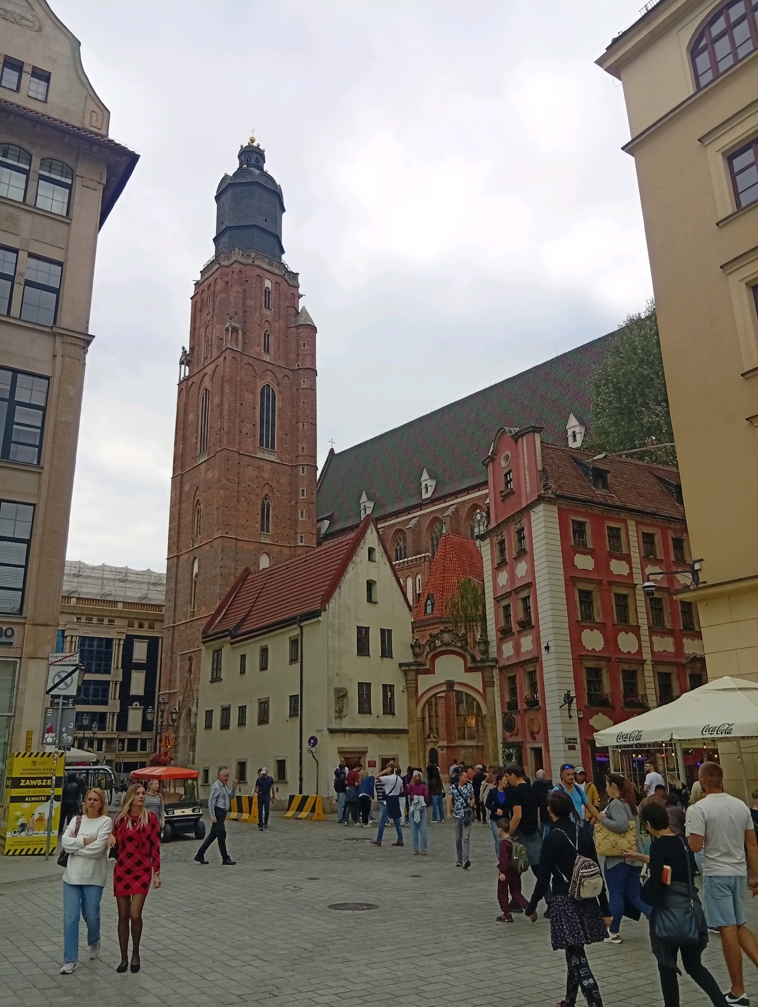 This is a medium-shot, eye-level, exterior, daytime photograph of a public square in Wrocław, Poland, featuring the iconic St. Elizabeth's Church and surrounding historical buildings. The sky is overcast.
In the foreground, two women are walking towards the viewer. The woman on the left has blonde hair, is wearing a white sweater and blue jeans, and is carrying a dark handbag. The woman on the right has blonde hair, is wearing a red and black checkered dress, black stockings, and black heels. She is looking forward.
Behind them, the cobblestone square is populated with a diverse group of people. Some are walking, others are standing and conversing. A small orange electric vehicle is parked to the left of the center. A large yellow warning sign with black stripes and Polish text is visible on the far left.
The prominent architectural feature is St. Elizabeth's Church, a large, red-brick Gothic tower with a dark green spire. It is flanked by other historical buildings with red tile roofs. To the left of the church, a modern building with large windows is visible. To the right, a light yellow building with red trim and arched windows stands next to a bright pink building with white decorative elements. A white Coca-Cola branded umbrella is set up to the right, suggesting outdoor seating for a cafe or restaurant.
The overall scene depicts a lively urban square, likely a popular tourist destination given the presence of the church and the mix of locals and visitors.