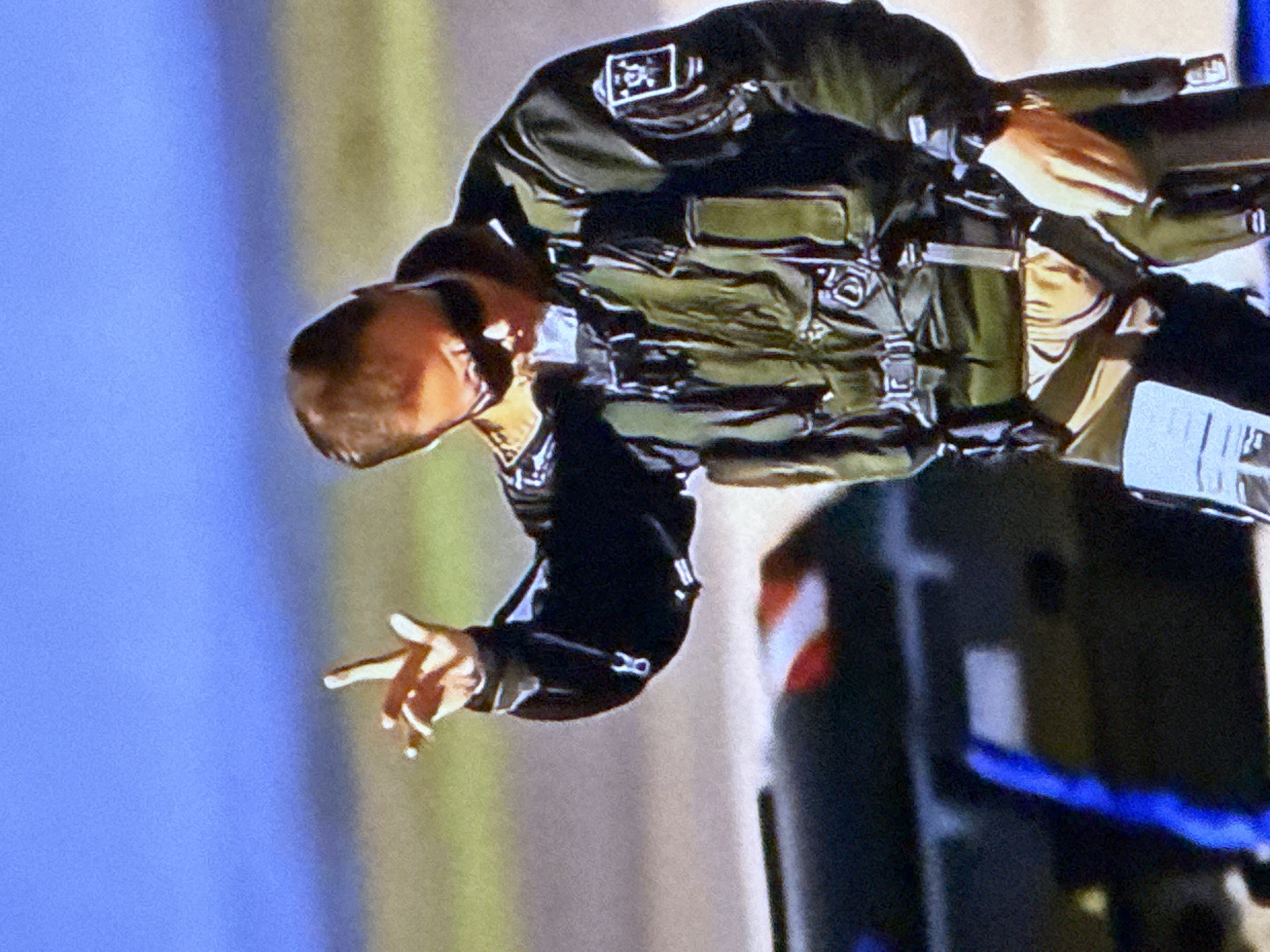 The image shows a person in a green flight suit with a patch on the left sleeve, wearing sunglasses. The person is standing outdoors with a blue sky in the background. A dark object, possibly a vehicle or aircraft, is visible in the lower left portion of the image. The person is making a hand gesture with their index and middle fingers raised. There is no information in the image to suggest a location in the city of Cahul, Moldova.