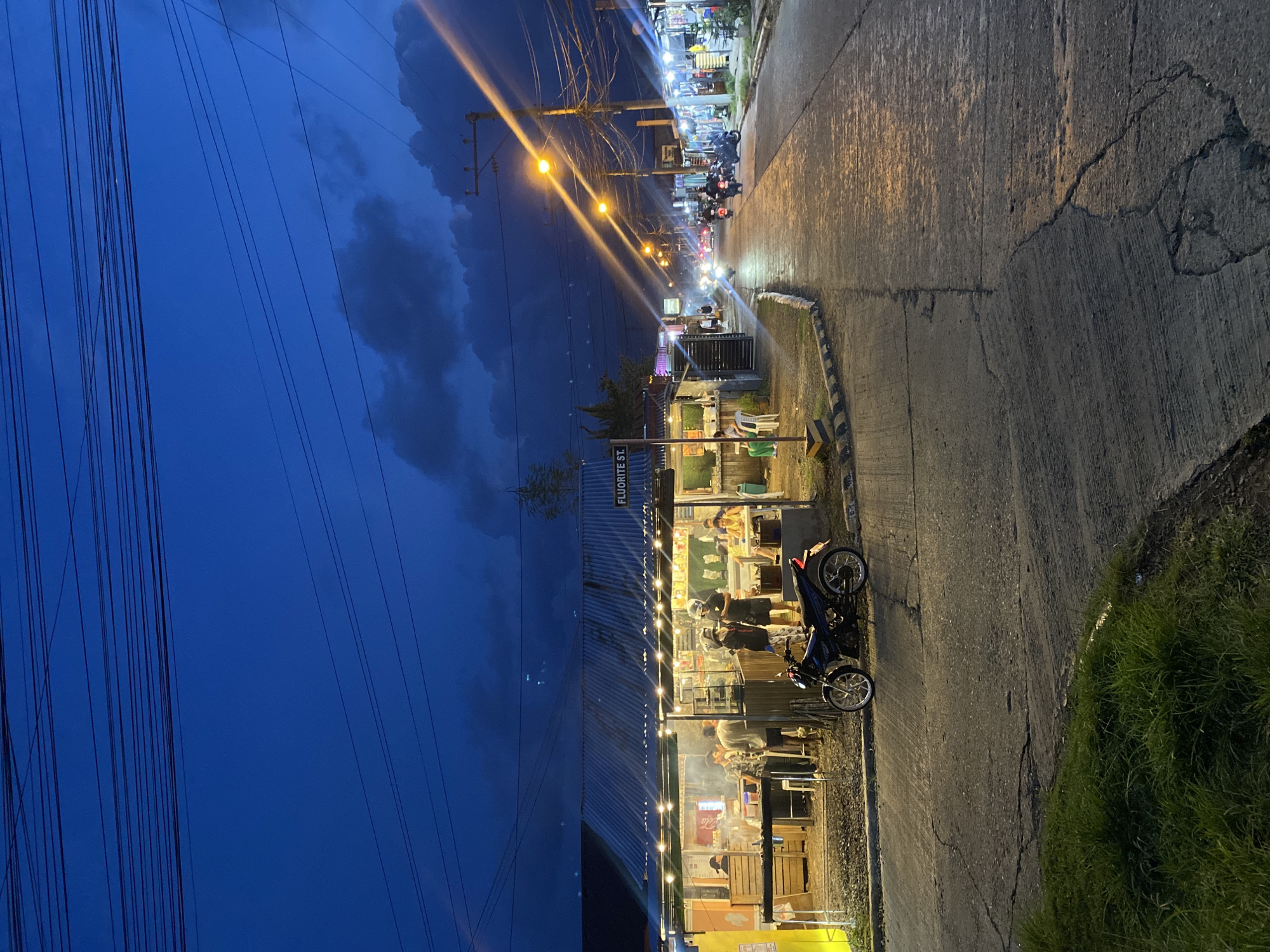 This image depicts a street scene at dusk in Davao City, Philippines. The sky is a deep blue, transitioning from day to night, with scattered dark clouds. Overhead, a dense network of electrical wires crisscrosses the sky.
On the left side of the image, a row of small establishments with corrugated metal roofs are visible, illuminated by string lights. People are gathered in front of these stalls, appearing to be customers or vendors. One establishment has a sign that reads "FLUORITE ST.". A parked motorcycle is positioned in front of these stalls.
The street itself is paved and appears to be well-lit by streetlights, which cast long streaks of light across the asphalt. Several motorcycles are seen on the street, some in motion and some parked. The right side of the street is lined with more businesses, also illuminated by lights. The overall atmosphere suggests a bustling street market or commercial area in the evening.