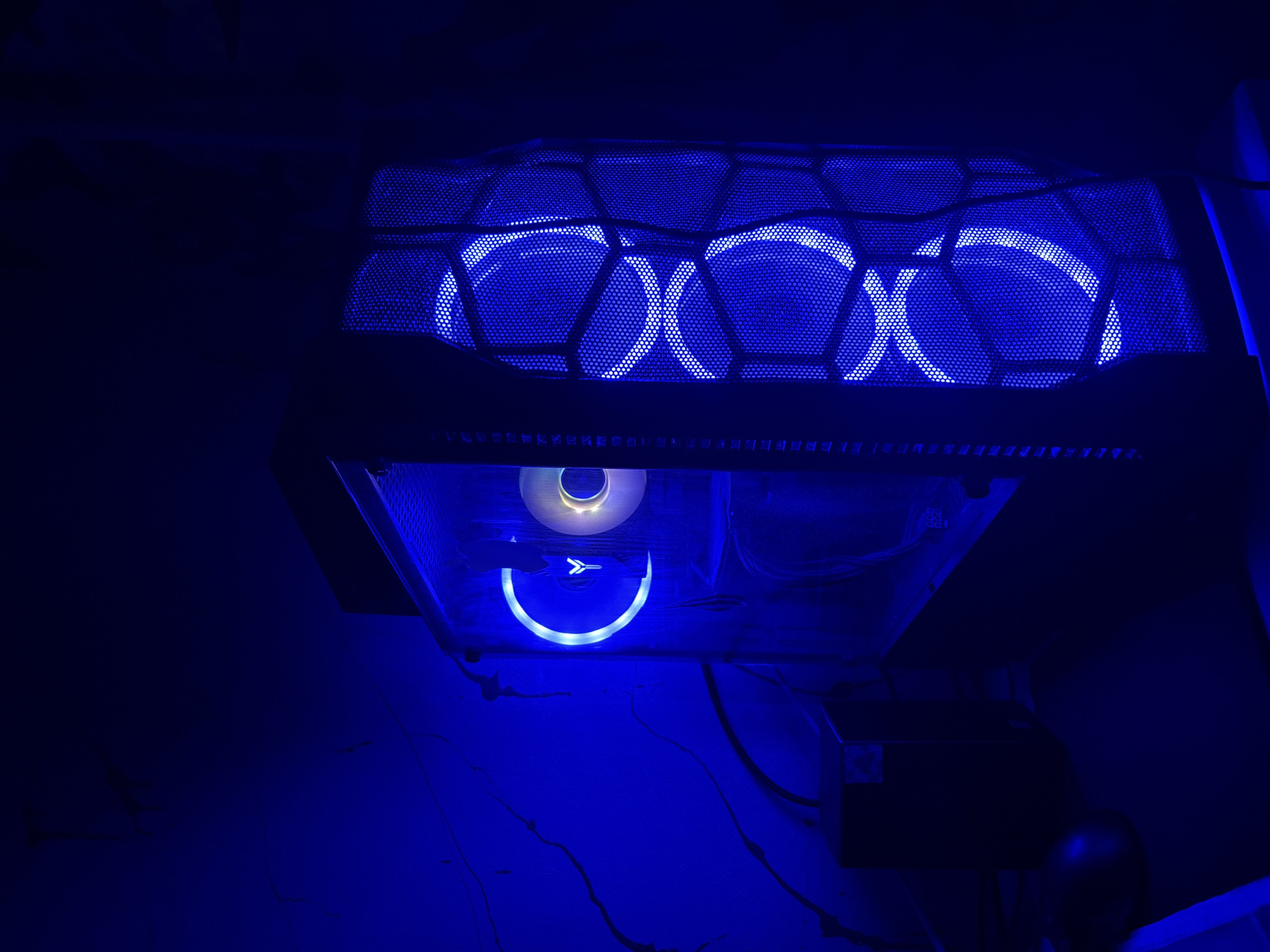 The image shows a computer tower with blue LED lighting. The top of the tower has a mesh grill with three fans visible underneath, each with a circular blue light ring. Below the top, the side of the tower appears to be made of glass, revealing some internal components. A circular fan with a blue light ring and a white logo is also visible inside the tower. The overall scene is dark, with the blue light from the computer being the primary illumination. There is no visible text in the image. The setting is likely indoors, given the presence of the computer equipment. The lighting creates a futuristic or gaming aesthetic.