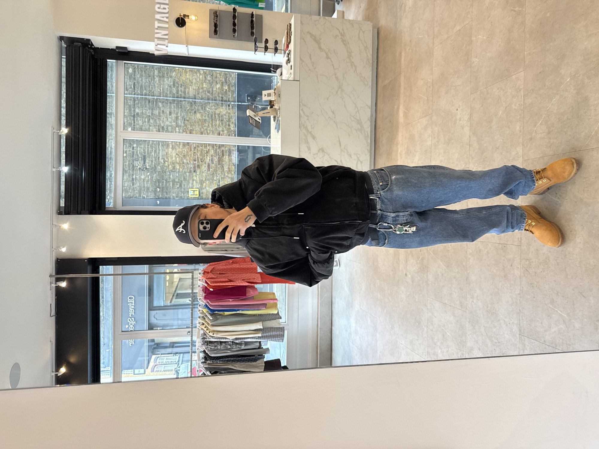 A person is seen taking a photo of themselves in the mirror. The individual is wearing a black jacket, blue jeans, and tan boots. The reflection shows a storefront with a "Vintage" sign and clothing racks. The location appears to be a shop, judging by the clothing on display and the general commercial setting. Outside the shop window is an urban street scene. Without additional elements, it's impossible to accurately identify the specific location in the city of London.