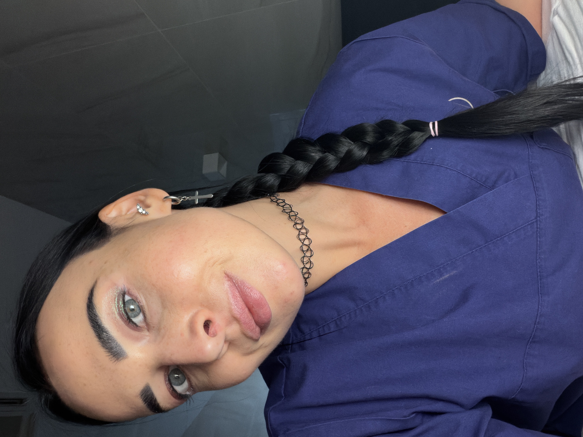 The image is a close-up, rotated portrait of a young woman, likely a selfie. Her head is tilted significantly to her right (viewer's left), making her appear to look directly into the camera from a slightly upward angle. The background is indoor, featuring a dark, indistinct wall or ceiling on the left and a lighter, possibly striped or textured white/grey surface on the right.
The woman has fair skin with subtle freckles and a somewhat dewy complexion. Her features include dark, well-defined eyebrows and striking light-colored eyes, possibly blue or grey, enhanced with shimmery light green/silver eyeshadow, dark eyeliner, and mascara. Her lips are a soft pink with a slightly pouting expression. Her long, dark hair is styled in a thick, neatly woven single plait that rests over her right shoulder and chest area, secured near the end with two pink elastic bands.
She is dressed in a dark blue garment that resembles medical scrubs or a professional uniform. Her visible left ear is adorned with multiple pieces of jewelry: a small, sparkling stud with three stones near the helix and a silver hoop earring from which a small cross dangles. Around her neck, she wears a black choker necklace with an intricate, openwork, web-like pattern.
Her expression is engaging and direct, conveying a neutral yet confident demeanor. There is no visible text in the image. This media file is associated with the location context of Saint Petersburg, Russia.