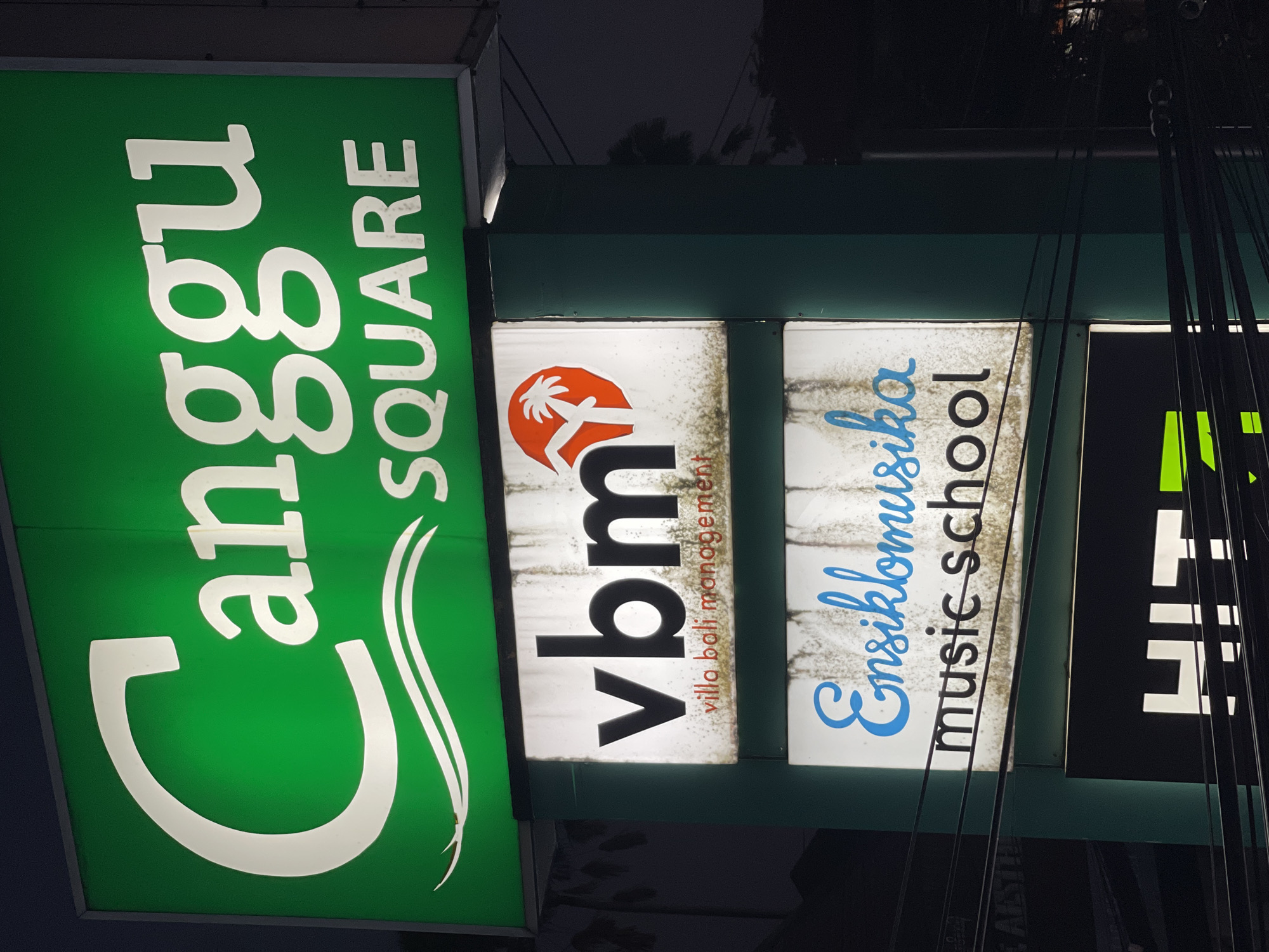 The image shows a vertical sign in North Kuta, Indonesia. The topmost sign is green with white letters spelling out "Canggu SQUARE." Below that is a sign for "vbm villa bali management" in black letters with an orange palm tree symbol. Next is a sign that reads "Ensiklomusika music school" in blue and black letters, respectively. A final sign, mostly obscured by power lines, has bright green letters on a black background. The overall lighting is dim.