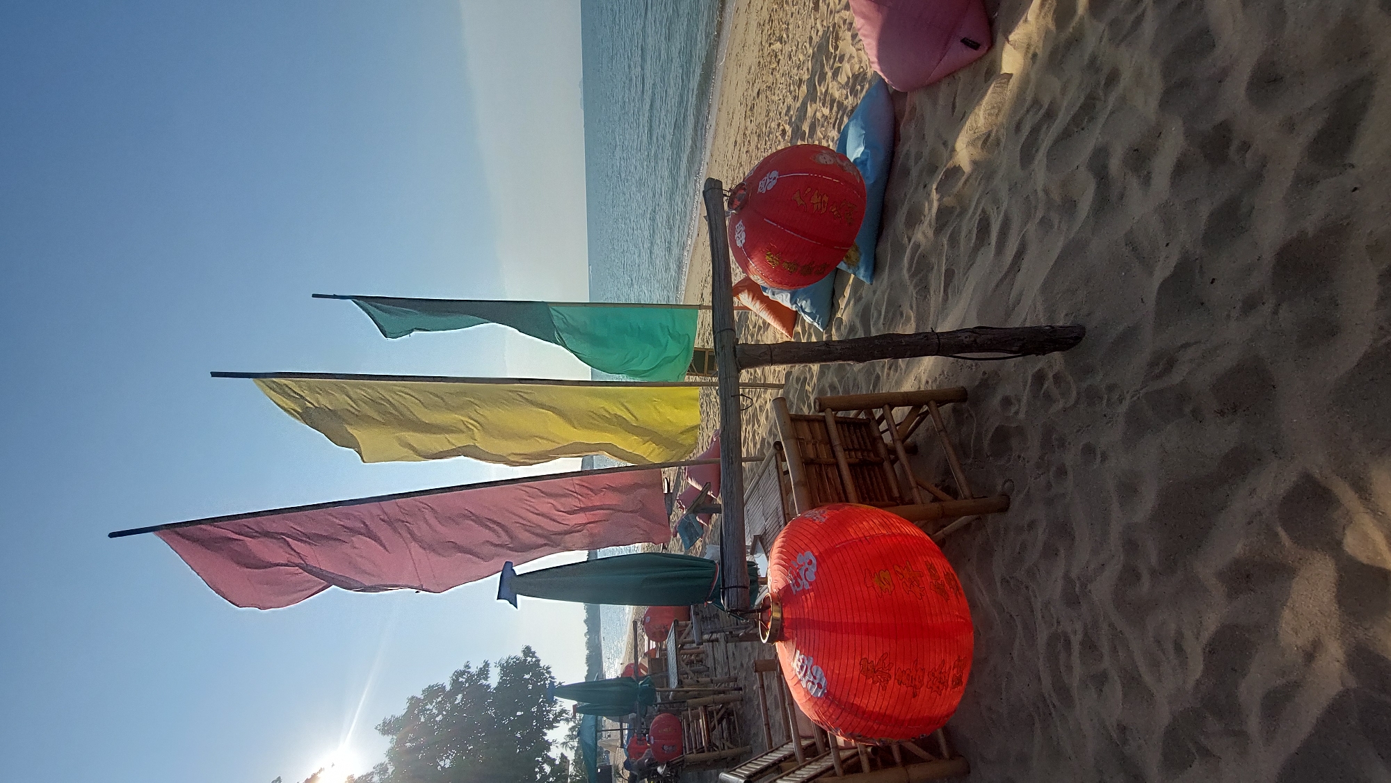 The image, captured in Baan Mae Nam, Thailand, presents a serene and colorful beach setting, likely during the late afternoon. The scene is dominated by a clear blue sky, visible across the upper portion, with the sun casting a bright flare from the bottom-left, partially obscured by distant foliage. Long shadows stretch across the sandy ground, indicating the low angle of the sun.
In the midground, several vibrant decorative flags, including teal, yellow, and pink, are strung on poles, waving gently. A rustic wooden post, holding some of these flags, also supports two prominent red, spherical paper lanterns. These lanterns are adorned with intricate white patterns, possibly stylized characters or motifs. More lanterns are visible further back along the beach.
The foreground and midground feature light-colored sand, showing subtle textures and footprints. Scattered on the sand are elements of a casual beach lounge area, including traditional bamboo chairs and tables. Near the water's edge, soft, comfortable seating in the form of pink, light blue, and orange cushions or beanbags invites relaxation. The calm, blue-green ocean stretches across the right side of the image, meeting the sandy shore and extending to the horizon.
While no people are visible, the arrangement of the decorative flags, lanterns, and seating suggests a peaceful and inviting atmosphere for leisure or dining, characteristic of a beachside resort, cafe, or relaxation zone. The overall impression is one of tranquility and tropical charm. No clearly legible text is visible, only decorative patterns on the lanterns.