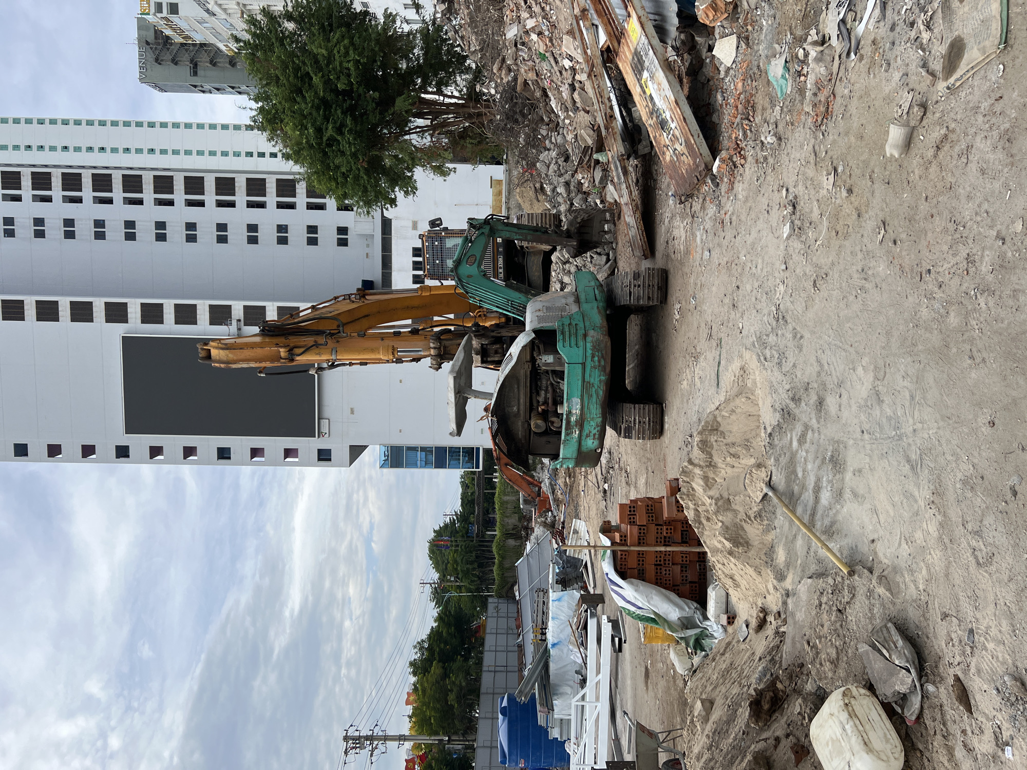 The image depicts a construction or demolition site in Nha Trang, Vietnam, with two excavators as the central focus. One excavator, predominantly green and teal, is positioned in the foreground, its engine compartment open, suggesting it is either undergoing maintenance or has been damaged. Its bucket is lowered towards the ground. Behind it, a larger yellow excavator arm is visible, extending upwards and to the left, possibly belonging to another machine or the same one from a different angle.
The site is cluttered with debris, including piles of bricks, rubble, metal sheeting, and a large blue water tank. A mound of sand is present in the foreground, with a shovel resting against it. Several white metal structures are also scattered around the site, some appearing to be scaffolding or construction supports.
In the background, towering modern buildings rise against a cloudy sky. One prominent building is a white high-rise with numerous rectangular windows and a large dark rectangular screen or advertisement panel. To the right, another building displays signage that includes "VENUE" and "BAY CITY." The overall environment is dusty and appears to be under active construction or renovation, typical of urban development in Nha Trang. No people are visible in the image.