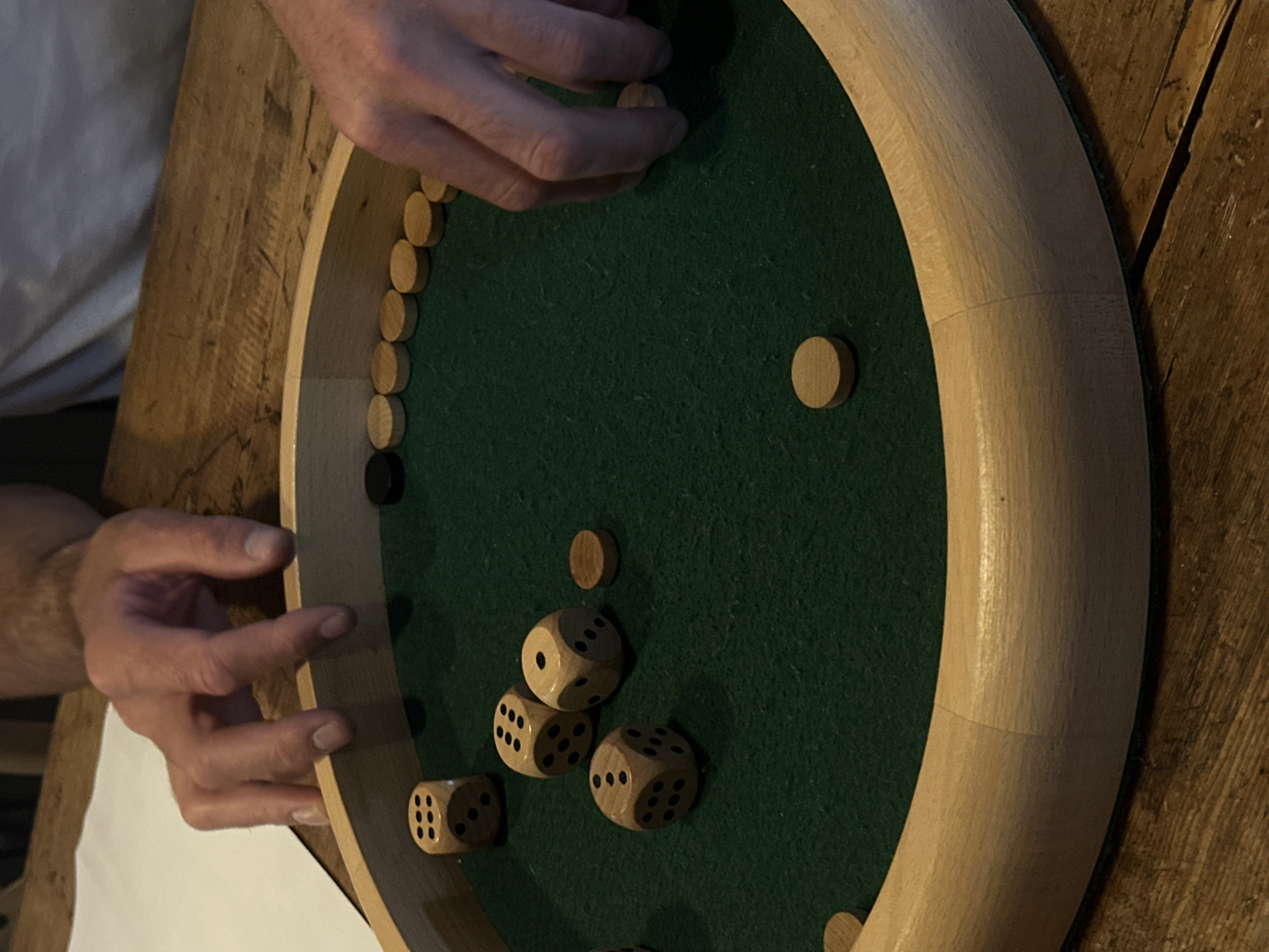 A person is playing a game on a circular wooden board with a green felt surface. The game involves wooden discs, some light and one dark, arranged around the edge of the board and scattered in the center, along with several dice. The person's hands are visible as they manipulate the discs. The board rests on a wooden surface, and a piece of white paper is visible on the left side of the image. The location is not evident from the image, and therefore cannot be associated with La Couarde-sur-Mer, France.