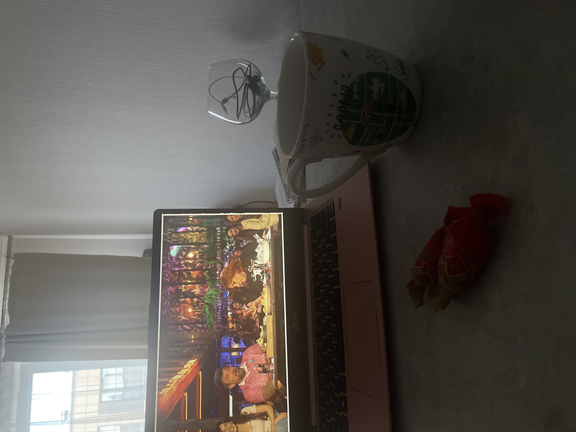 An indoor scene, likely in Poroshkino, Russia, shows a laptop open on a table, displaying a television show or video. The laptop screen shows several people seated at a table, appearing to be engaged in a conversation or discussion. To the right of the laptop, a white mug with a design on it is positioned on the table. Above the mug, a wine glass contains black wires. Two red candies with gold accents are placed in the foreground, slightly out of focus. A window with light-colored curtains is visible in the background, suggesting daytime. The lighting in the room appears to be a mix of natural light from the window and possibly artificial light. The overall ambiance suggests a relaxed, casual setting.