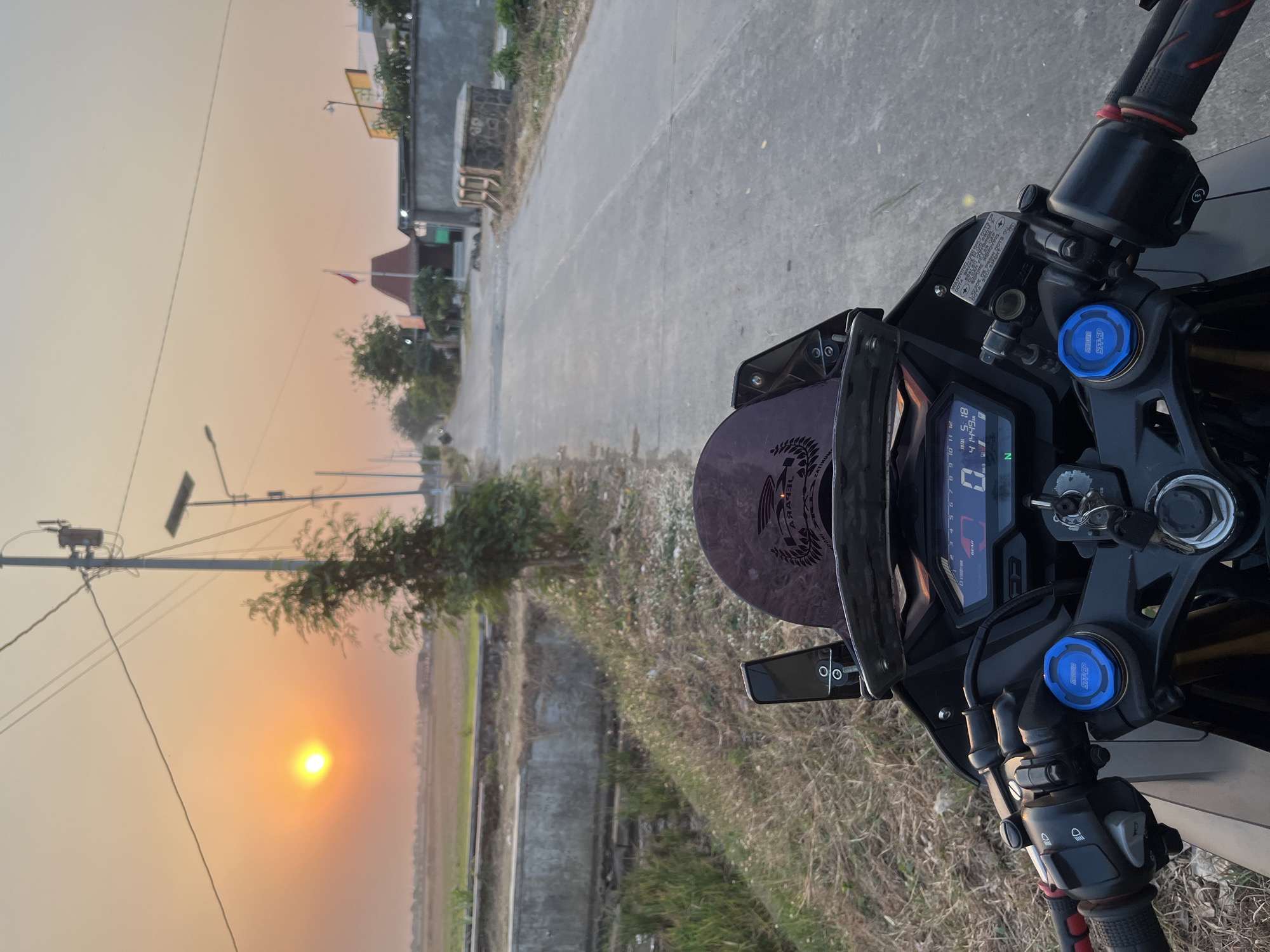 The image depicts a first-person view from a motorcycle on a paved road in Pecangaan, Indonesia, during sunset. The sun is visible in the upper left quadrant, casting a warm, orange glow across the sky and horizon. The road stretches forward, flanked by dry grass and a concrete structure on the left, and a grey wall and some buildings on the right. A few trees are scattered along the roadside. In the foreground, the motorcycle's handlebars, dashboard, and a portion of its fairing are visible. The dashboard displays digital readouts, including speed and time. A dark, tinted windscreen is mounted above the dashboard. A red and white flag flies from a building in the distance on the right side of the road. Power lines and streetlights are also present. The overall scene suggests a quiet, rural or semi-rural setting at dusk.