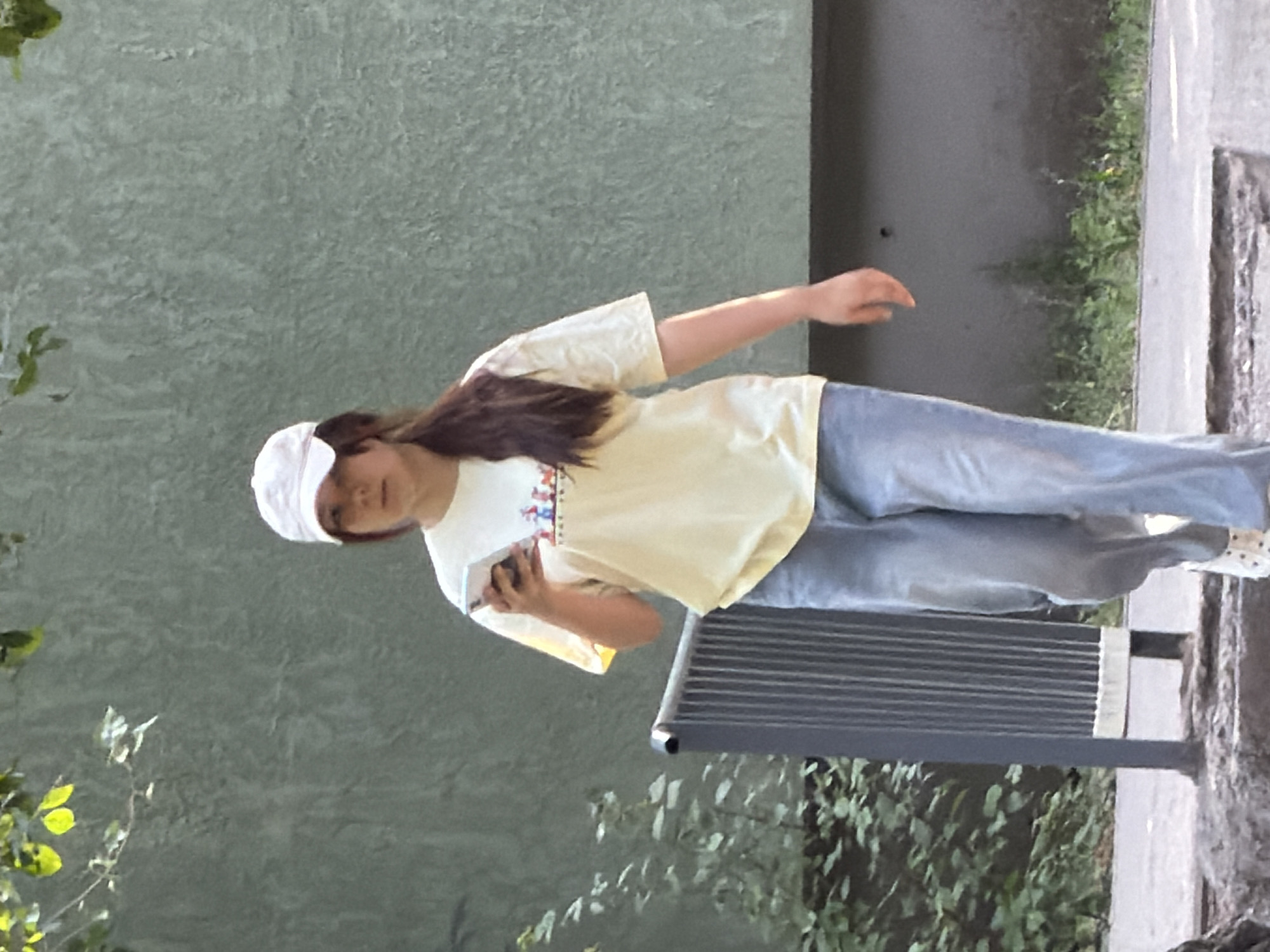 A young woman with long, dark hair pulled back and wearing a white baseball cap, a light yellow t-shirt with a graphic print, and light blue jeans walks towards the viewer while looking at her smartphone. She is in an outdoor urban environment, with a textured, light green wall behind her. To her left, a dark grey metal trash receptacle with vertical slats stands beside some foliage. The ground in front of her appears to be a paved walkway, with a curb and some rougher ground with debris. The lighting suggests it is daytime.
