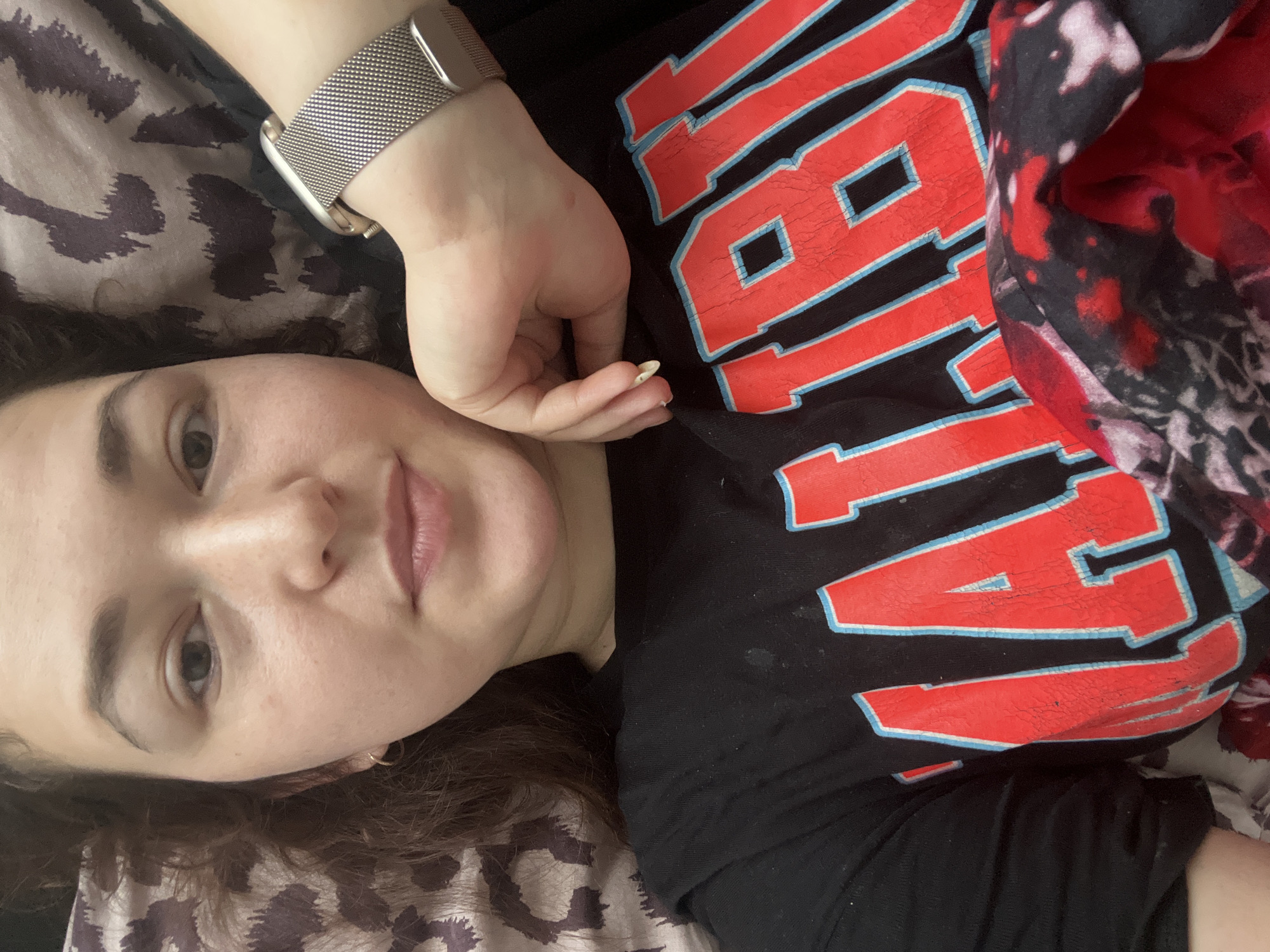 The image shows a close-up, overhead view of a young woman lying down and looking at the camera. She has brown curly hair, fair skin, and is wearing a black t-shirt with red and blue lettering that appears to say "MALIBU". A silver watch is visible on her left wrist. The background consists of patterned bedding. There is no discernible information in the image that would allow for an identification of the location within Chelyabinsk, Russia.
