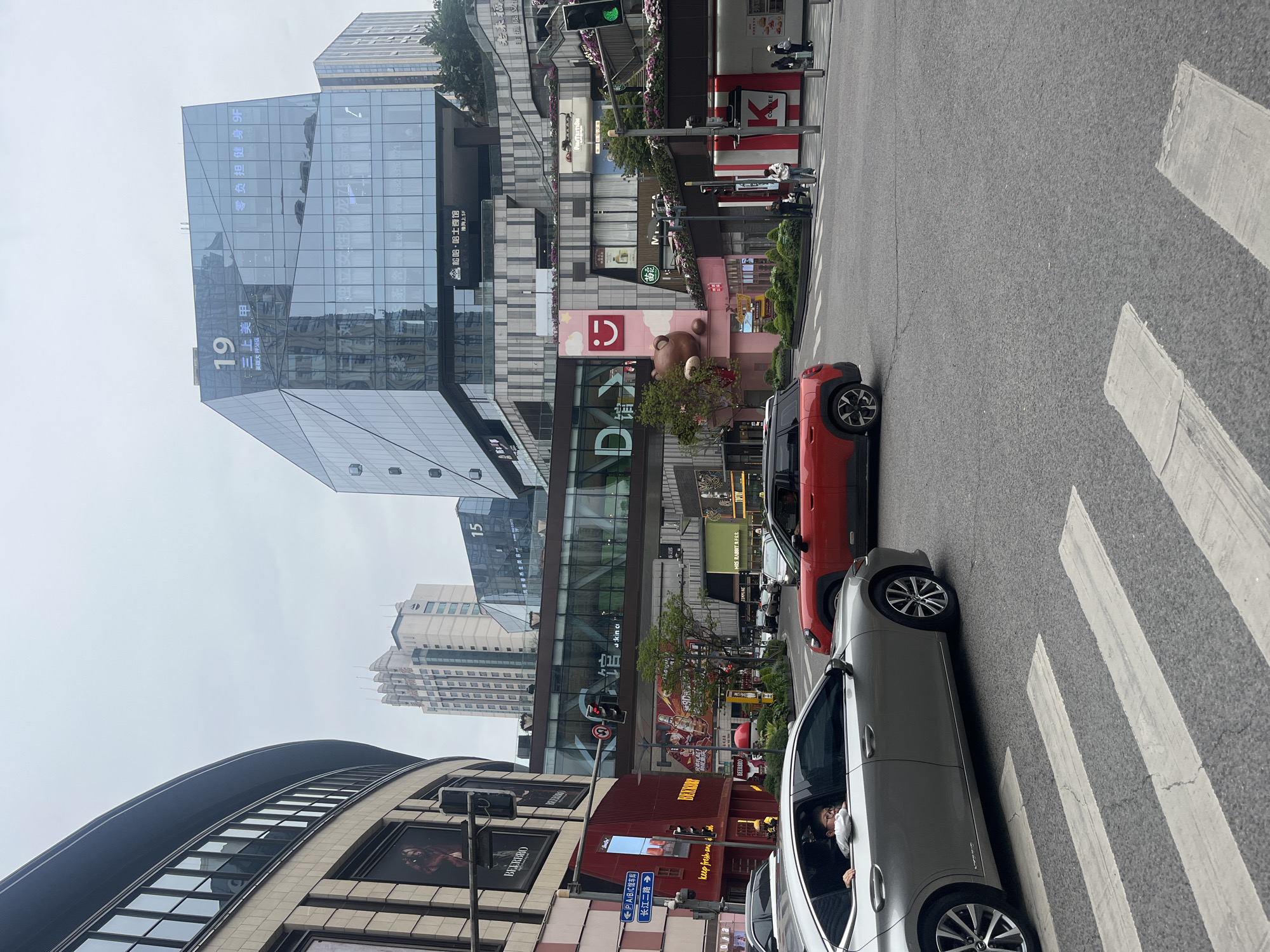 The image depicts a street scene in the Yuzhong District of China. Prominent architectural features include a curved building with "BEERBRO" signage, a modern glass building labeled "19 三上美甲," and a structure with a large teddy bear image. Two cars, one silver and one red, are situated on a zebra crossing. A pedestrian is visible in the silver car. Several signs and storefronts line the street, including "BEERBRO" and "keep fresh ans." Additional buildings are visible in the background. The sky appears overcast. The street is marked with crosswalks. Traffic lights are positioned above the street.