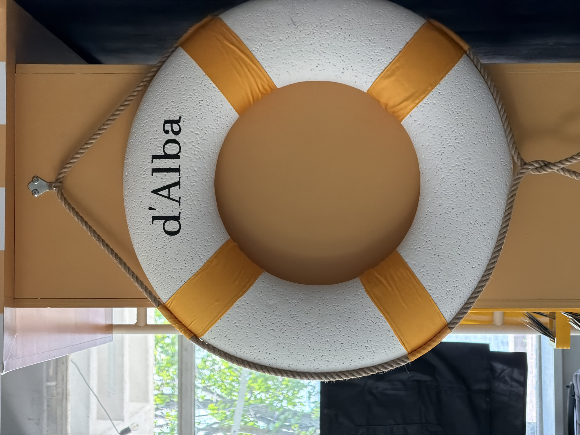 A white lifebuoy, textured to resemble a rough surface, with four uniformly spaced orange fabric bands, is depicted prominently. A braided natural fiber rope borders the outer edge of the lifebuoy and forms a loop at the top, suspending it from a silver-toned metal hook. The hook, secured by two visible screws, is affixed to a smooth, matte orange vertical surface, likely a wall or pillar. The name "d'Alba" is inscribed in black serif letters on the upper half of the lifebuoy.
To the left of the lifebuoy, through a window, lush green foliage from outdoor trees or bushes is visible, suggesting a natural environment outside. Portions of a light-colored building or structure, possibly including steps or a railing, are partially visible beyond the foliage. A single bare light bulb on a wire hangs in the upper left corner of the frame. In the lower left foreground, a dark, draped object, possibly fabric or a garment, partially obscures the view through the window. To the right of the orange pillar, a dark, uniform vertical surface is visible, indicating an adjacent wall or architectural feature. The scene is an interior view, specifically identified as the d'Alba restaurant located at Aptekarskaya Embankment 20, Saint Petersburg, Russia. No individuals are present in the image.
