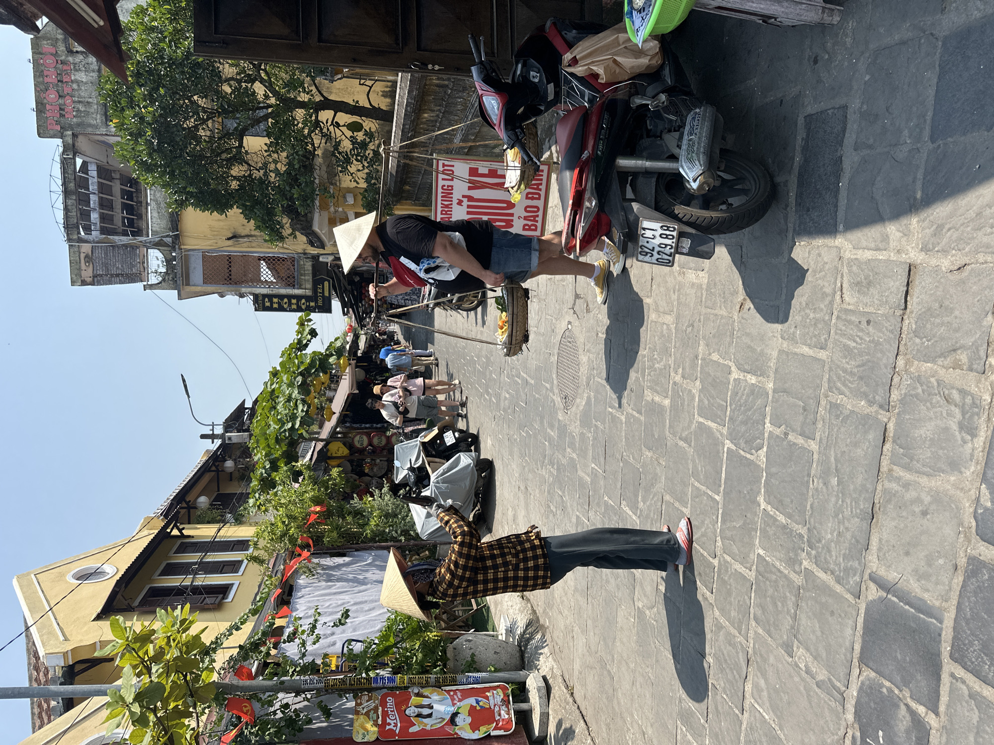 The image shows a street scene in Hội An Ward, Vietnam, with two individuals wearing traditional Vietnamese conical hats ("nón lá"). One person, dressed in a brown and black checkered shirt, is standing near a red advertisement for "Merino" ice cream. The other person, in a black t-shirt and shorts, is near a parked red and black motorcycle. A sign for "Parking Lot" and "Pho Hoi Hotel" are also visible, as well as a license plate on the motorcycle that reads "92-C1 029.88." The street is paved with gray stone blocks. Other people are visible in the background. Buildings on both sides of the street exhibit Vietnamese architectural styles with yellow facades.