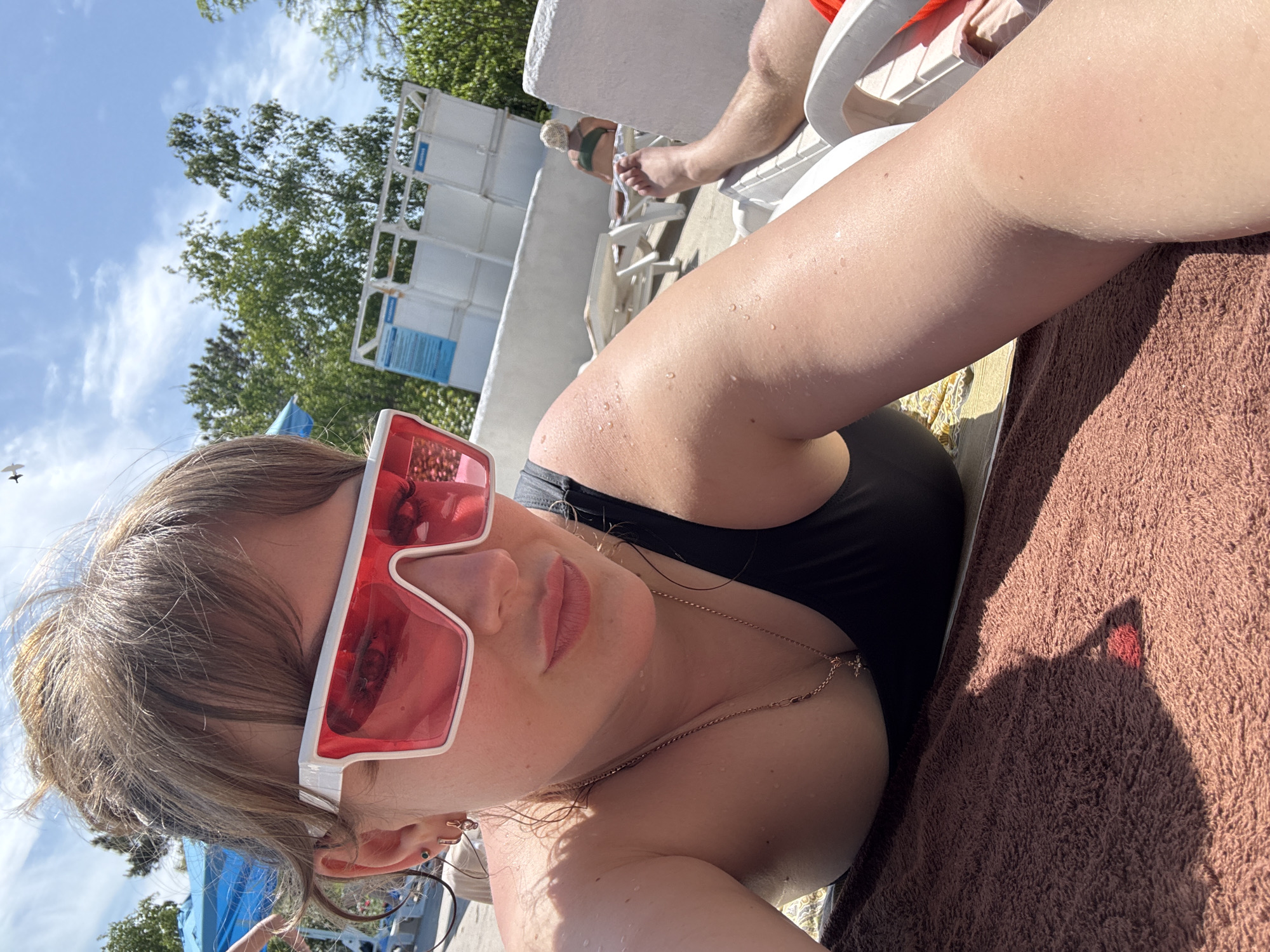 The image depicts a woman wearing pink-tinted sunglasses and a black swimsuit lying on a brown towel, possibly by a swimming pool. She is wearing jewelry, including a necklace. In the background, there are trees, a light blue sky, and a white structure that appears to be a building or a wall, possibly part of a pool facility. There are other people present, including a person lying on a lounge chair behind her. The location is possibly in Yevpatoriya, Ukraine.