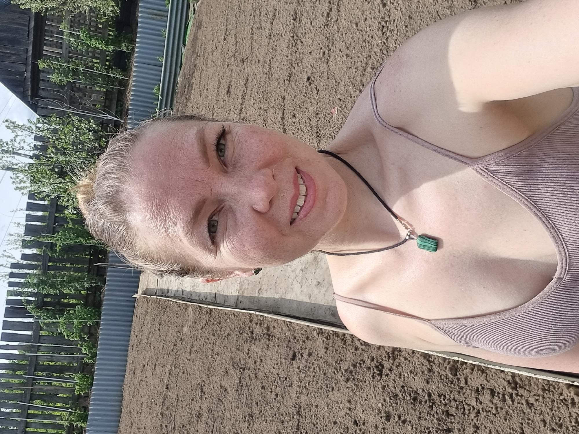 A woman with fair skin and light-colored hair pulled back in a bun is smiling at the camera. She is wearing a light purple tank top and a necklace with a green, rectangular pendant. She appears to be outdoors, with a cultivated plot of brown soil visible behind her. In the background, there is a wooden fence, some trees, and corrugated metal fencing. Based on the vegetation, it is likely a rural or suburban area. The image may have been taken in Chaikovsky, Russia.