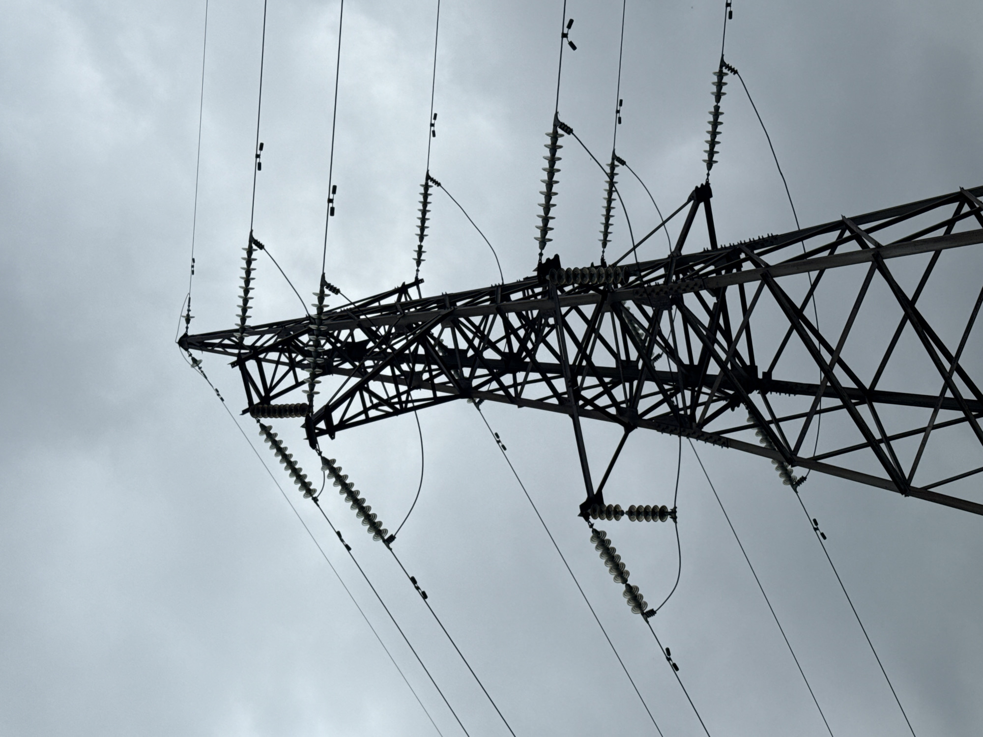 A power transmission tower stands against a cloudy sky in Kamensk-Uralsky, Russia. The tower is constructed of dark metal lattice and supports multiple power lines suspended by ceramic insulators. The lines are taut and extend across the frame. The sky is overcast, with a uniform grey tone. There are no people or other discernible objects or actions in the image.