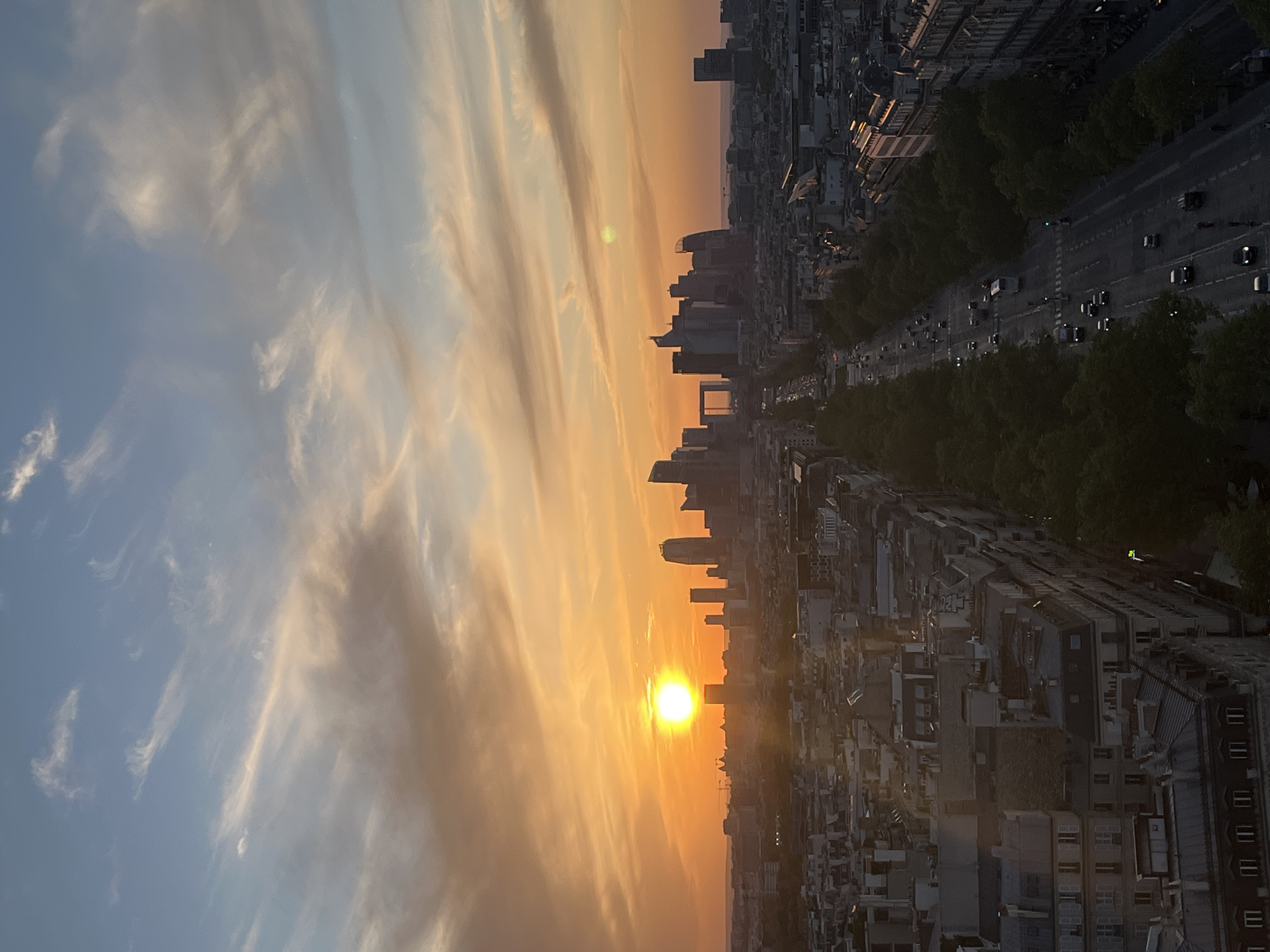This image captures a panoramic view of a city during either sunset or sunrise, creating a striking golden hour scene. The sky occupies a significant portion of the frame, transitioning from a clear blue in the upper parts to vibrant oranges, yellows, and amber closer to the horizon, where the bright sun is prominently featured. Wispy, cirrus-like clouds are scattered across the sky, catching the warm light and appearing in soft shades of white, pink, and orange.
Below the colorful sky, a dense urban landscape unfolds. In the foreground and midground, traditional multi-story buildings with pitched roofs and numerous windows line the streets, characteristic of Parisian architecture. A wide, tree-lined boulevard runs through the scene, filled with vehicles whose headlights and taillights are visible, indicating active traffic. The trees appear dark and lush against the fading light.
In the distance, a modern cityscape with taller buildings and skyscrapers is silhouetted against the brilliant sky. A distinct, large rectangular arch-like structure can be identified among these buildings, strongly resembling La Grande Arche de la Défense. This detail, combined with the general architectural style, confirms the location as Paris, France, likely viewed from an elevated position looking west towards La Défense during sunset.
The time of day is clearly either sunset or sunrise, given the low position of the sun and the warm, soft light bathing the scene. The weather appears fair with scattered clouds. There are no people visibly identifiable, nor is there any discernible text within the image. The overall mood is serene and picturesque.