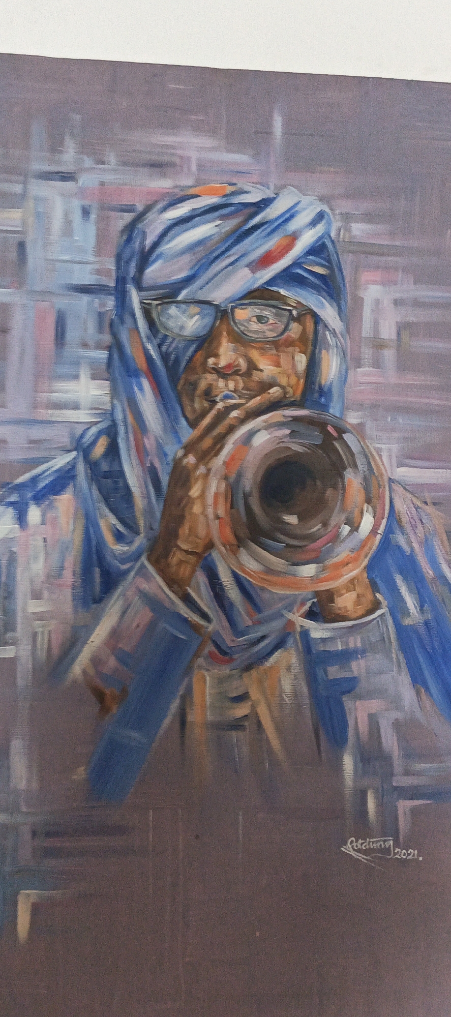 An oil painting depicts a dark-skinned individual, likely male, wearing eyeglasses and a blue and white striped head covering, reminiscent of a turban or tagelmust. The figure holds a brass instrument, resembling a trumpet or horn, to their mouth, indicating a musical performance. The background is abstract, composed of visible, broad brushstrokes in shades of grey, purple, blue, and light pink, creating an indistinct, possibly urban, environment. The art style is painterly and impressionistic. The person's gaze, discernible through the reflection in the eyeglasses, conveys focus and immersion in playing the instrument. In the lower right corner, the artist's signature "Rezdung" and the year "2021" are visible. The artwork, situated in the context of Akwanga, Nigeria, captures a moment of musical expression.
