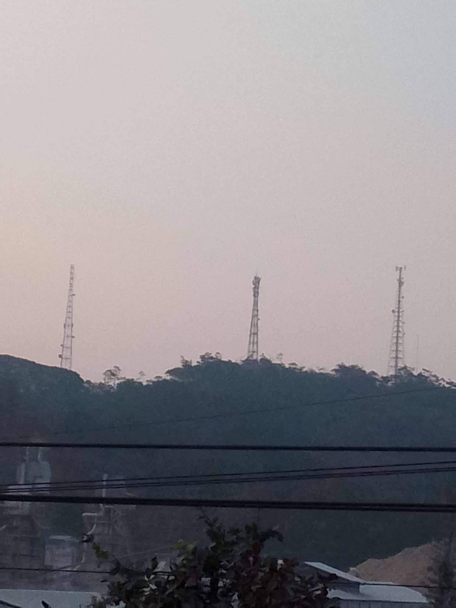 The image shows a hazy, overcast sky over a landscape dominated by a tree-covered hill. Three tall, lattice-like towers, presumably communication or transmission towers, stand on the crest of the hill, spaced apart. In the foreground, several dark power lines stretch horizontally across the frame. Below the hill and partially obscured by the power lines, structures that appear to be part of a building or complex, including what looks like a large statue, are visible. The overall scene suggests an outdoor environment, possibly on the outskirts of a town or city, with a focus on the elevated terrain and the man-made structures upon it. The location is identified as Gunungmasigit, Indonesia.