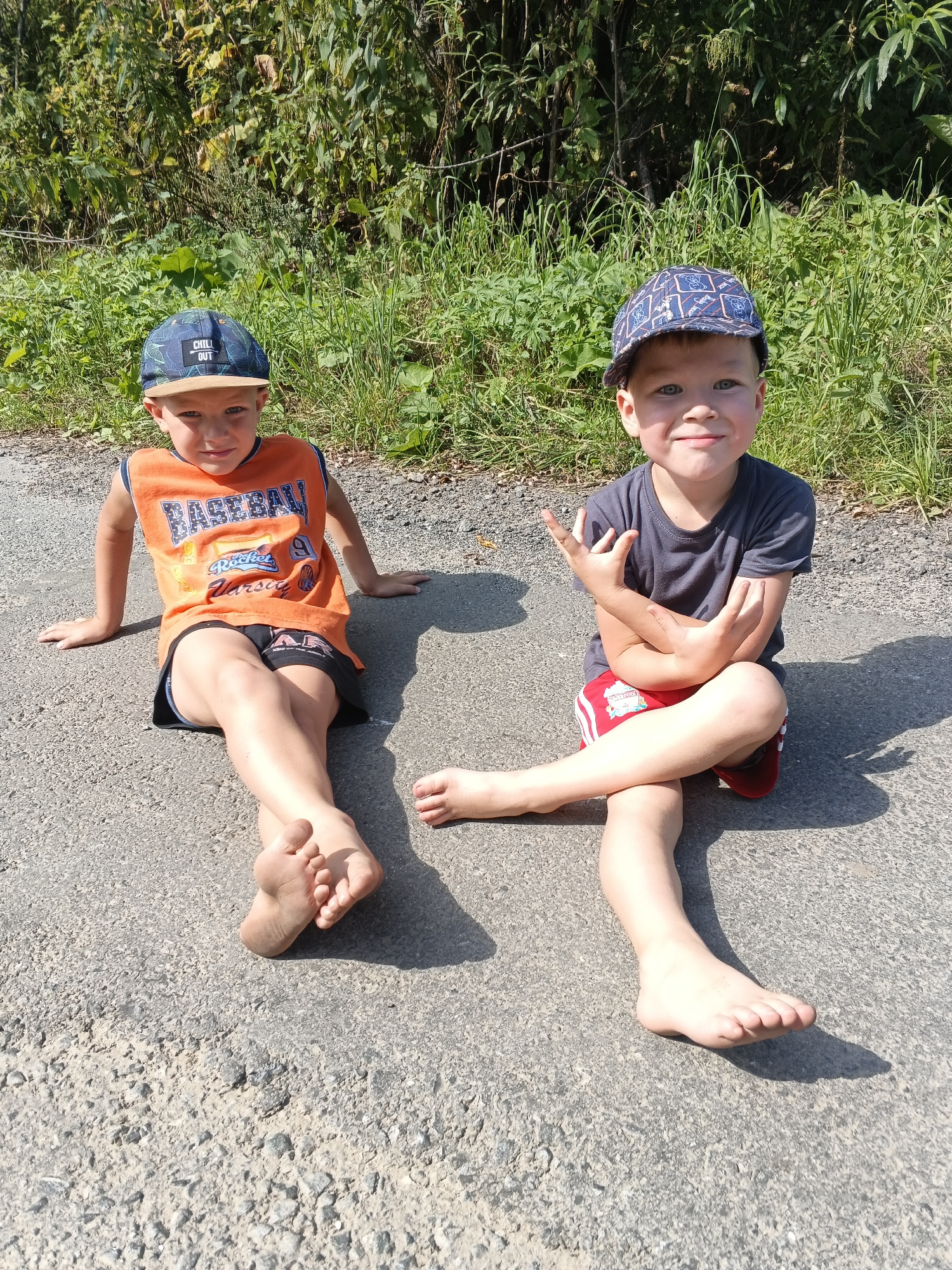 Two young boys are sitting on a paved surface, possibly a road or path, in what appears to be a rural or semi-rural outdoor setting. The ground is asphalt with visible gravel. Behind them, there is a dense area of green vegetation, likely bushes and trees, with some dry leaves scattered on the ground. The lighting suggests it is daytime, with distinct shadows cast by the subjects.
The boy on the left is wearing an orange sleeveless t-shirt with "BASEBALL" and "Rocket Varsity" graphics, and dark shorts. He has a blue baseball cap with a tan brim and "CHILL OUT" text. He is sitting with his legs extended, one foot slightly raised, and his hands resting on the ground behind him.
The boy on the right is wearing a dark grey t-shirt and red shorts with white stripes. He also has a blue baseball cap with a patterned design. He is sitting with his legs crossed and his arms folded, with his right hand making a "rock on" gesture. His left leg is extended forward. Both boys are barefoot.
The setting suggests a casual, outdoor activity, possibly a break during a walk or a visit to the countryside. The specific location in Urtiushka, Russia, is not evident from the visual information alone, but the natural environment and the paved surface are consistent with many outdoor areas in such locations.