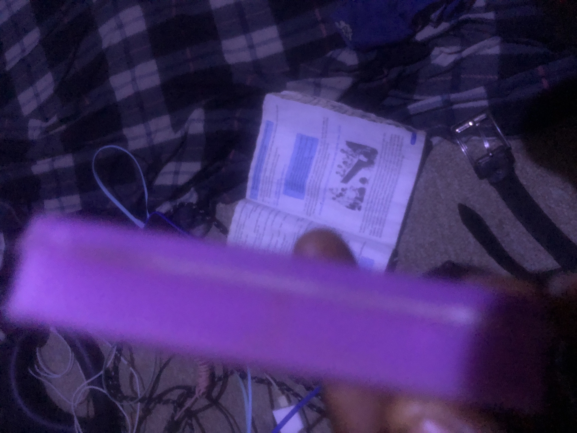 A close-up, low-light, blurry indoor shot shows a person's hand holding a rectangular purple object in the foreground. In the background, an open book with text and an illustration of people is visible, along with a dark plaid blanket and various cables. The scene appears to be in Jalingo, Nigeria.