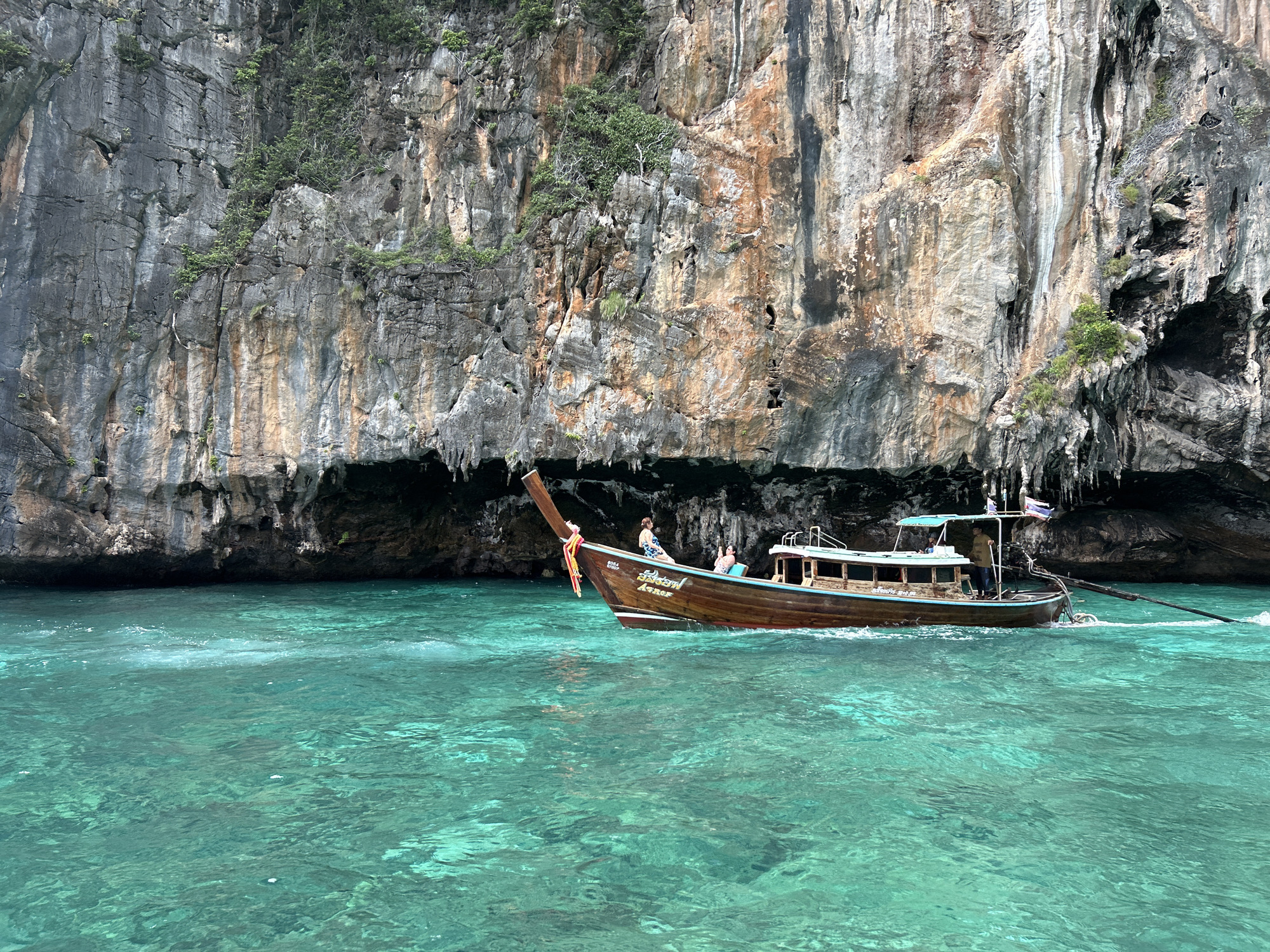 A long-tail boat, its wooden hull a rich brown, moves across turquoise waters. The boat has a sheltered seating area with a blue canopy and several people are visible. One person sits in the front, another stands near the back, and a third is seated under the canopy. A Thai flag is affixed to a pole at the rear of the boat. The boat's bow is adorned with decorative tassels of red and yellow. Towering limestone cliffs, sparsely vegetated, rise dramatically from the water's edge in the background, casting a dark shadow at their base. The water is clear and vibrant, with ripples indicating the boat's movement. The scene is consistent with the limestone karst topography characteristic of islands and coastal areas in Thailand, such as those found near Haad Yao.