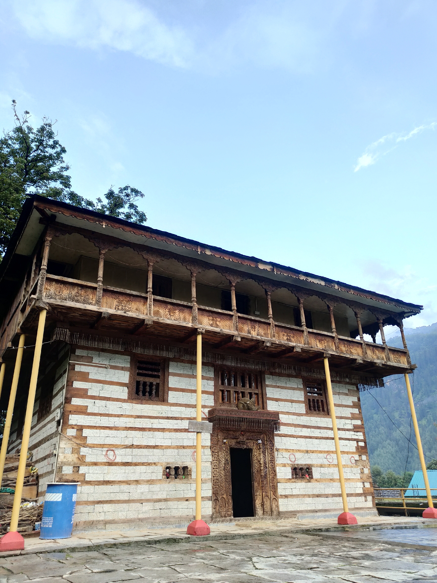 The image shows a traditional wooden and stone building, likely a temple or a historical dwelling, in Burwa, India. The building features multiple stories with ornate wooden balconies and intricately carved doorways and windows. The walls are constructed from alternating layers of stone and wood. Yellow support columns with red bases are visible, holding up the upper structure. In the foreground, a blue plastic barrel and some stacked wood are present, along with a stone-paved courtyard. The background reveals a lush green hillside and a cloudy sky. The scene appears to be an exterior shot, captured during daylight.