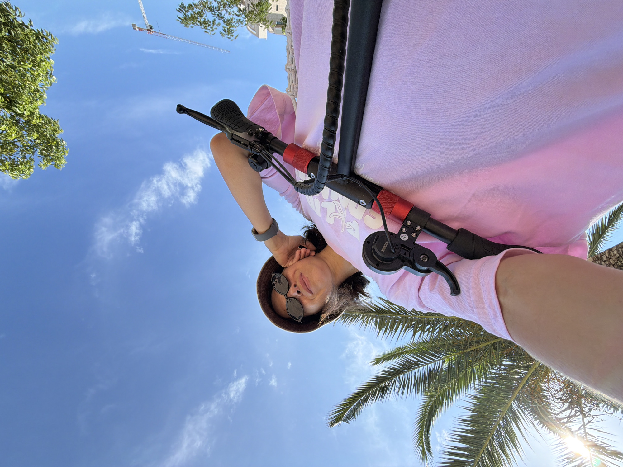 A person is captured from a low angle, looking up at a clear blue sky with scattered clouds. The individual is wearing a light pink hoodie, sunglasses, and a brown bucket hat, obscuring their eyes. They are holding onto the handlebars of an electric scooter, with parts of the scooter, including red accents on the handlebars and black grips, clearly visible. The person's hand is resting on their head, and they appear relaxed or perhaps shielding themselves from the sun.
The scene is set outdoors, with lush green palm fronds in the foreground, suggesting a warm, sunny day. In the background, a construction crane is visible against the sky, hinting at ongoing development or building work. The bright sunlight and clear sky indicate it is daytime, likely late morning or afternoon. The overall mood conveyed is one of leisure and enjoyment of the outdoors. The context of Budva, Montenegro, places this scene in a coastal European setting, known for its sunny climate and popular tourist destinations. No legible text is visible in the image.