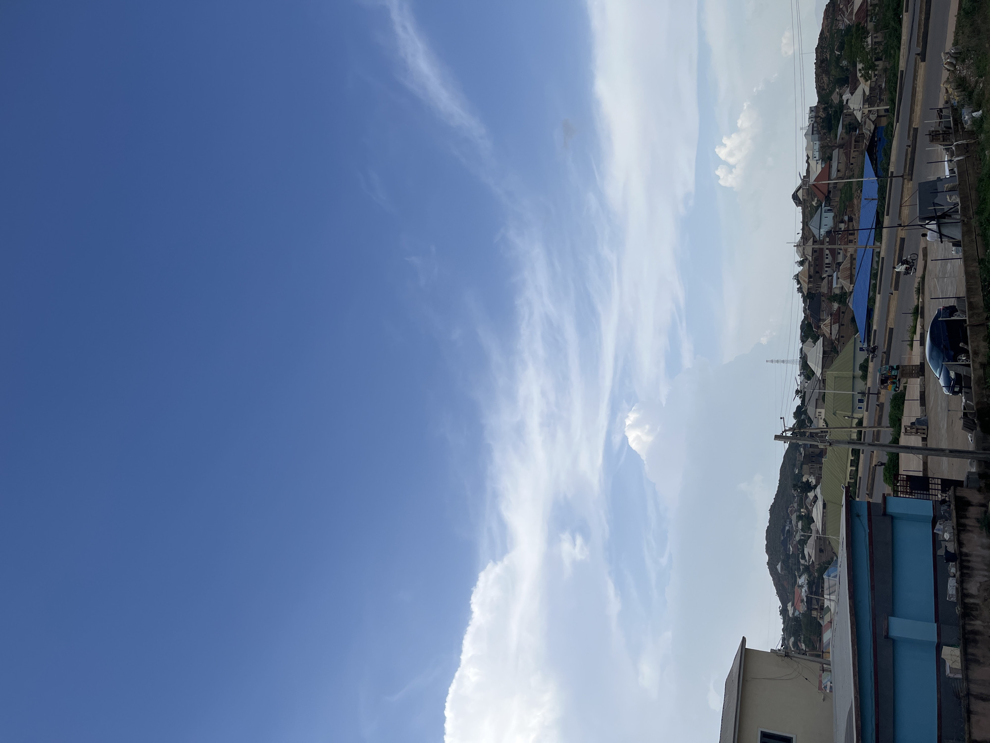 The image captures a daytime scene in the city of Gura Top, Nigeria. The sky is a vibrant blue, with scattered white clouds drifting across it. Below the sky, a dense residential area sprawls across a hilly terrain. Houses with corrugated metal roofs in various colors, including green, white, and blue, are densely packed together. Roads wind through the community, with a few cars and motorcycles visible, suggesting ongoing activity. The foreground features a portion of a building with a blue and grey facade. The overall impression is of a bustling urban environment nestled within a natural landscape.