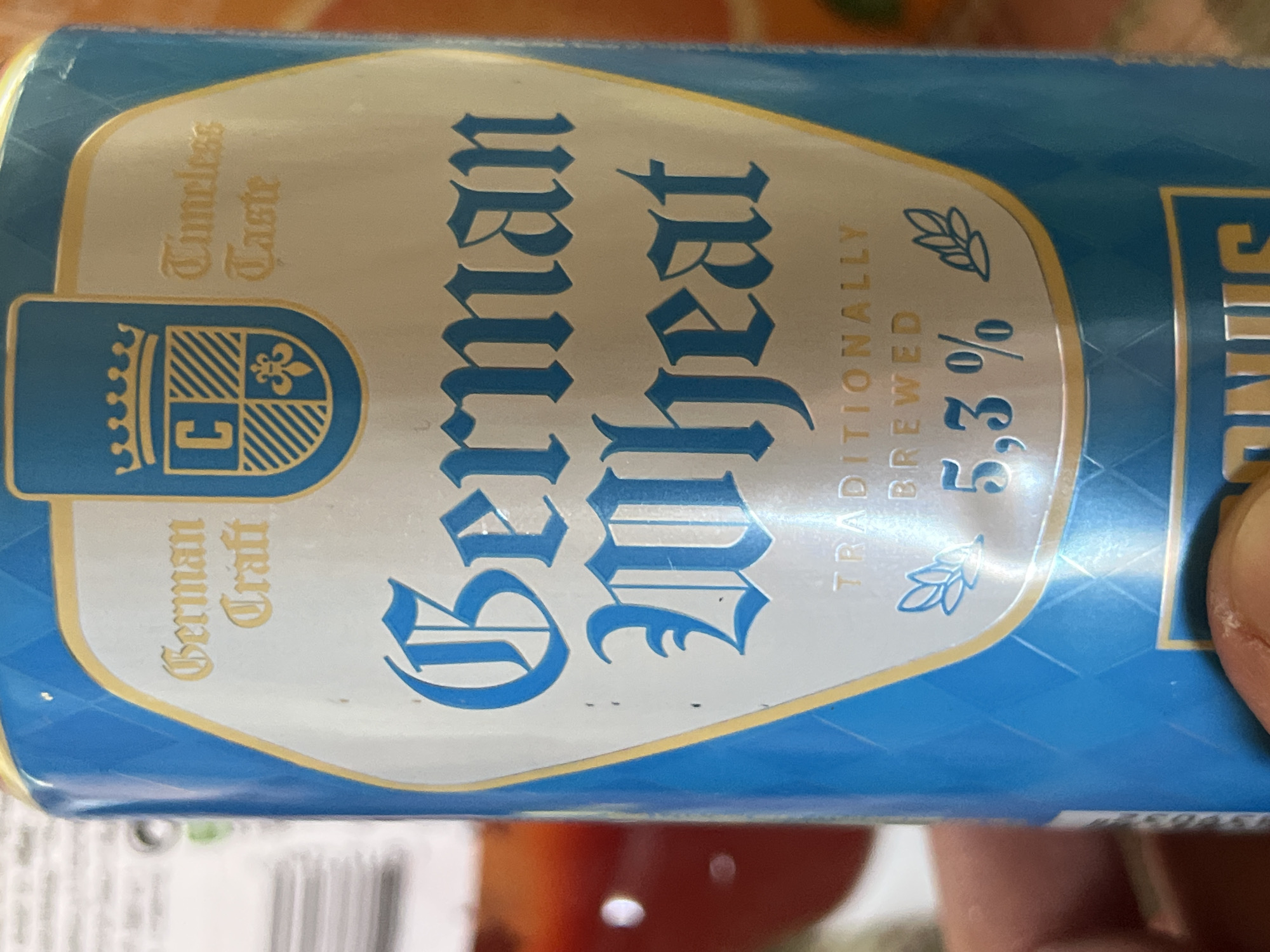 The image is a close-up, eye-level, indoor shot of a can of "German Wheat" beer. The can is predominantly blue with a white oval label that features gold lettering and a crest. The text on the label reads "German Craft," "Timeless Taste," "German Wheat," "TRADITIONALLY BREWED," and "5,3%". A stylized crest with a crown and a shield is also visible on the label. A finger is visible in the bottom foreground, holding the can. The background is blurred, showing what appears to be a table with some objects. There is no indication of the location being in Daulėnai, Lithuania.