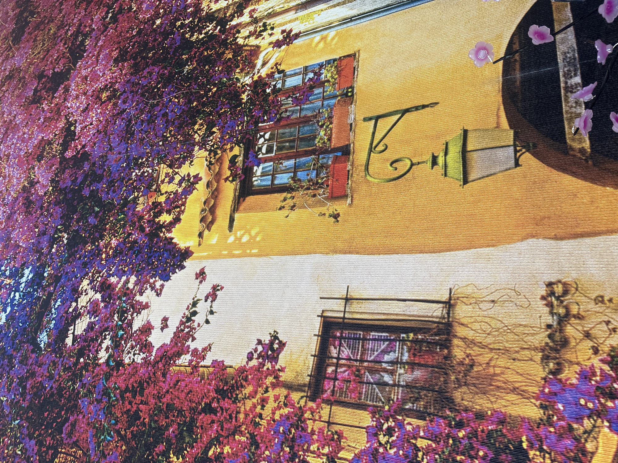 The image depicts a vibrant, sun-drenched facade of a building with a stucco finish in an ochre hue, possibly in the city of Georgiyevsk, Russia. Bougainvillea flowers, in shades of fuchsia, purple, and pink, cascade down from above, partially obscuring a window with a dark-colored frame and wrought-iron bars. The window itself has a multi-paned design and is adorned with red flower boxes containing green plants. Below this window, an ornate, green-painted lamp is mounted on a scrolling bracket. Further to the right, a portion of a dark, rounded object, possibly a doorway or archway, is visible, along with delicate pink floral decorations. The overall impression is one of Mediterranean charm or a stylized representation of such. There are no people or animals visible in the scene. The lighting suggests a bright, sunny day.