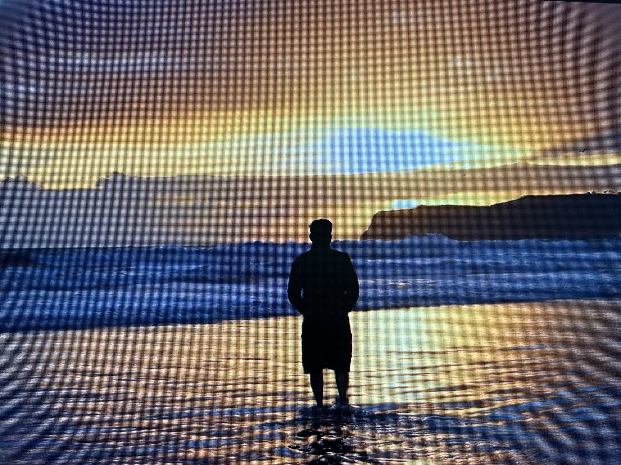 A solitary individual stands in the shallow water at the edge of a beach, facing a dramatic sunset over the sea. The sky is a vibrant tapestry of oranges, yellows, and blues, with clouds streaked across the horizon. Waves crash gently on the shore, reflecting the warm hues of the setting sun. The person, silhouetted against the bright sky, is dressed in shorts and a dark top. The water around their ankles shimmers with the reflected light. In the distance, a dark, rocky coastline is visible against the luminous sky. The scene evokes a sense of peace and reflection. This appears to be the coast of Varna, Bulgaria, given the characteristic landscape and the direction of the sunset over the Black Sea.