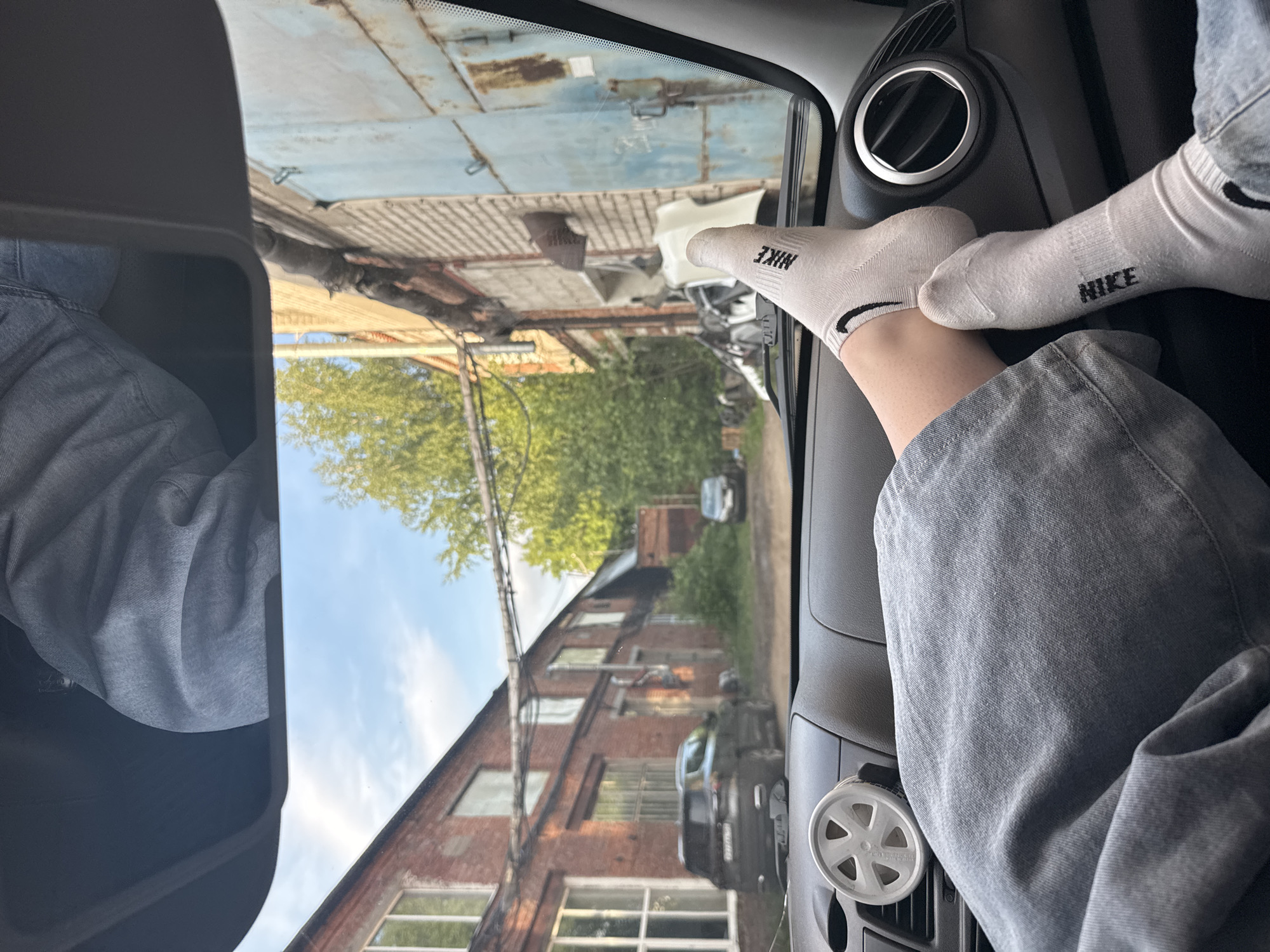A casual, low-angle shot taken from the passenger seat of a vehicle captures a relaxed moment with an urban industrial backdrop. The image is oriented vertically (originally rotated 90 degrees counter-clockwise).
In the foreground, a person's legs, clad in light-colored denim or jean material, are visible, with their feet resting on the dashboard or glove compartment area of the car. Both feet are covered in white low-cut socks, each prominently displaying the black "NIKE" logo. To the right, part of the car's interior dashboard is visible, featuring a circular air vent with a silver trim and a small, white, spoked air freshener or similar device. A rearview mirror is partially visible on the left side.
Through the car's windshield, an outdoor scene unfolds under a bright, clear sky. Directly outside, an old, weathered light blue metal corrugated building or garage door, streaked with rust, dominates the upper part of the exterior view. Below it, a cluttered area with various metallic debris and what appears to be discarded vehicle parts suggests an industrial yard or scrapyard. Further in the background, a multi-story red brick building with numerous windows indicates an older factory or warehouse structure. Green trees are visible between and behind these buildings, adding a touch of nature to the industrial setting. A dark colored car is partially visible parked near the brick building in the mid-ground. The scene suggests a location within an industrial or semi-industrial district, consistent with an urban area like Perm, Russia, during daytime with good weather.