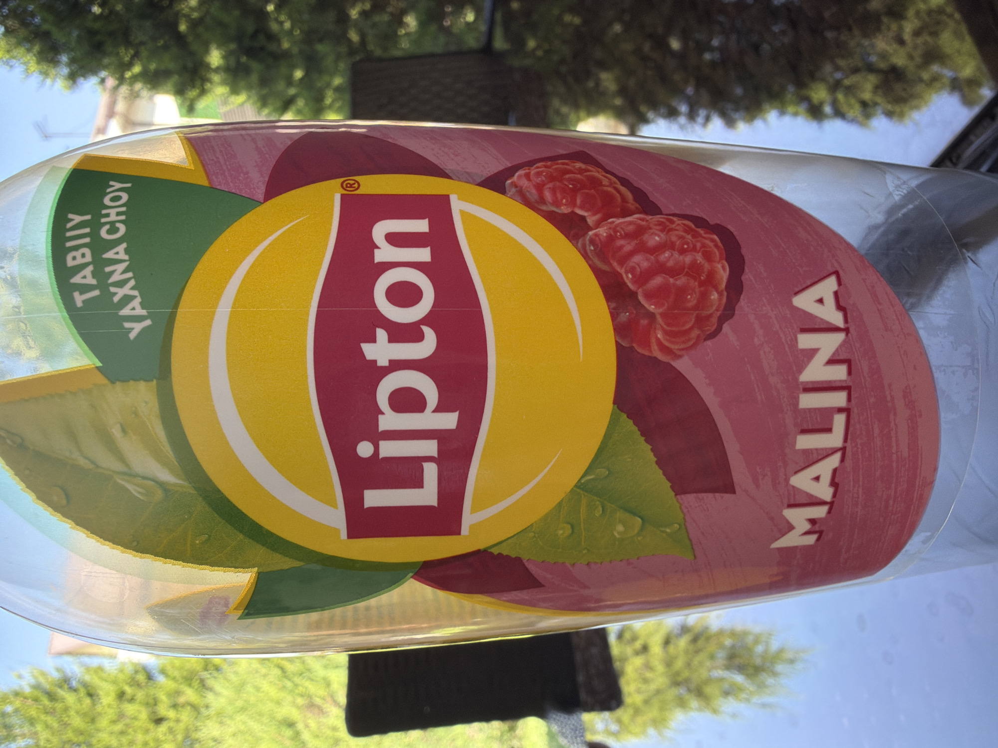 The image shows a close-up of a Lipton iced tea bottle with a raspberry flavor. The bottle is clear and plastic, with a label featuring the Lipton logo, green leaves, and images of raspberries. Text on the label includes "Lipton," "TABIIY YAXNA CHOY," and "MALINA." The bottle appears to be outdoors, possibly on a table, with trees and a blue sky visible in the background. The location of Koshkurgon, Uzbekistan, is not identifiable from the visual elements in the image.