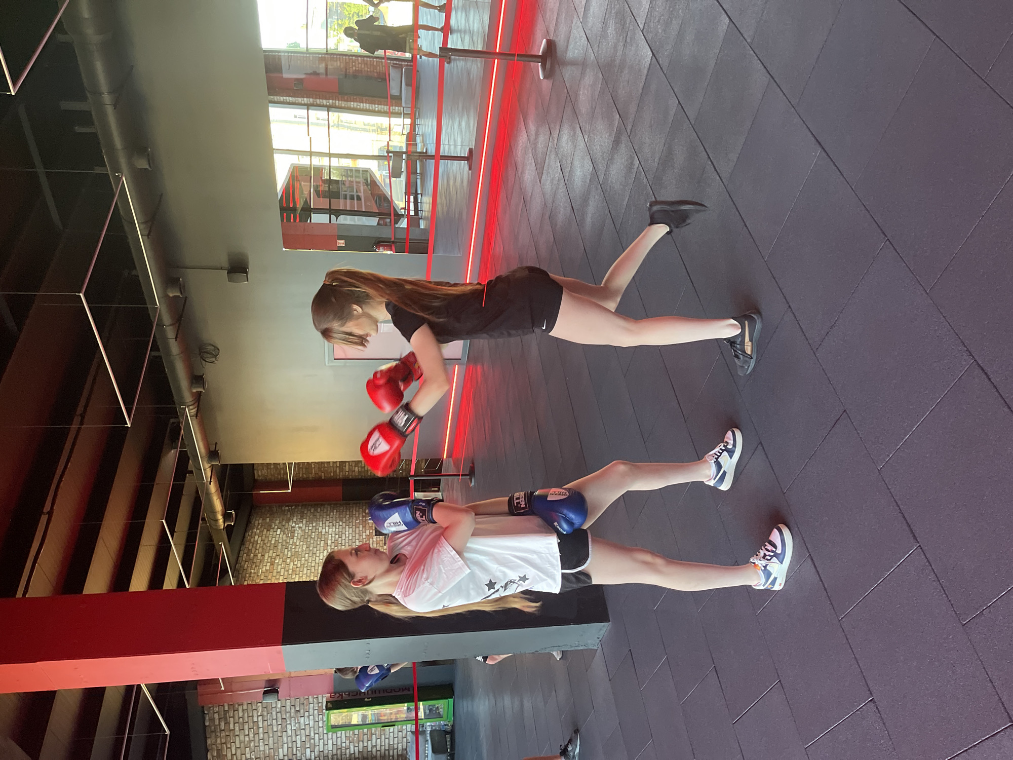 Two young women are engaged in a boxing training session in a gym. The woman in the foreground, wearing a white t-shirt with black stars and black shorts, has blue boxing gloves on. She is in a plank position with her hands on the floor. The woman behind her, wearing a black t-shirt and black shorts, has red boxing gloves on. She is also in a plank position, with one leg extended upwards. The gym floor is made of dark grey rubber tiles arranged in a diamond pattern. Red LED lights line the perimeter of a boxing ring in the background, with metallic poles supporting the structure. Large windows behind the ring offer a view of an outdoor area with greenery and a building. The overall lighting suggests it is daytime.