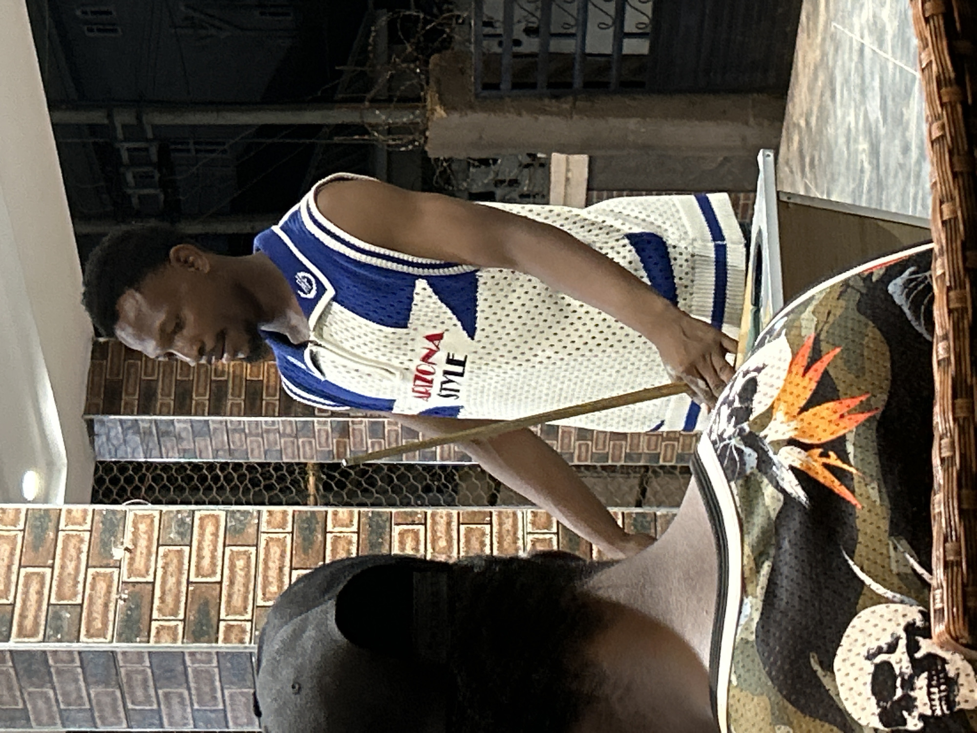 In Ihiagwa, Nigeria, an African man wearing a basketball jersey is playing pool with another man. The location appears to be outdoors, potentially a patio or semi-outdoor space with tiled walls.