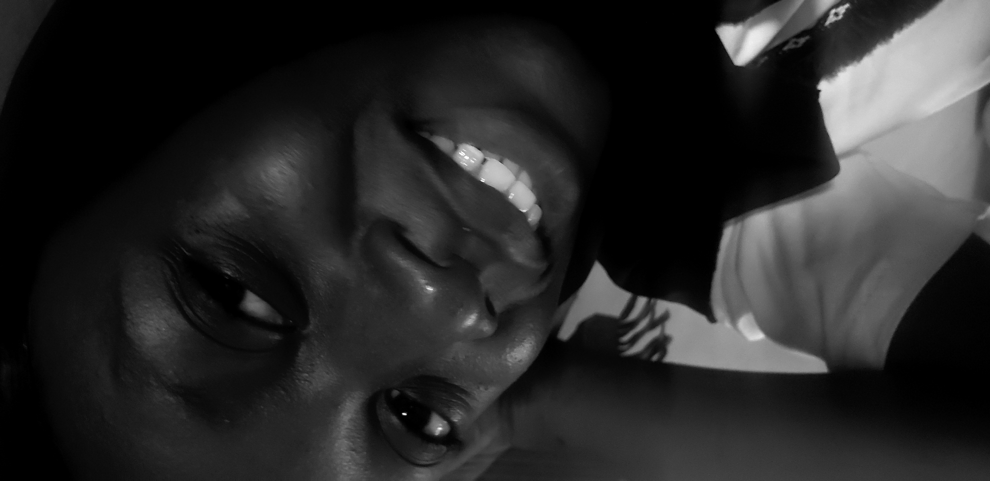 A monochrome close-up shows a person's face angled towards the viewer. The person is smiling, revealing teeth. Details of the face, including eyes, nose, and skin texture are visible. A light-colored shirt is partially visible on the right side. There is no indication of a specific location in Narayi, Nigeria.